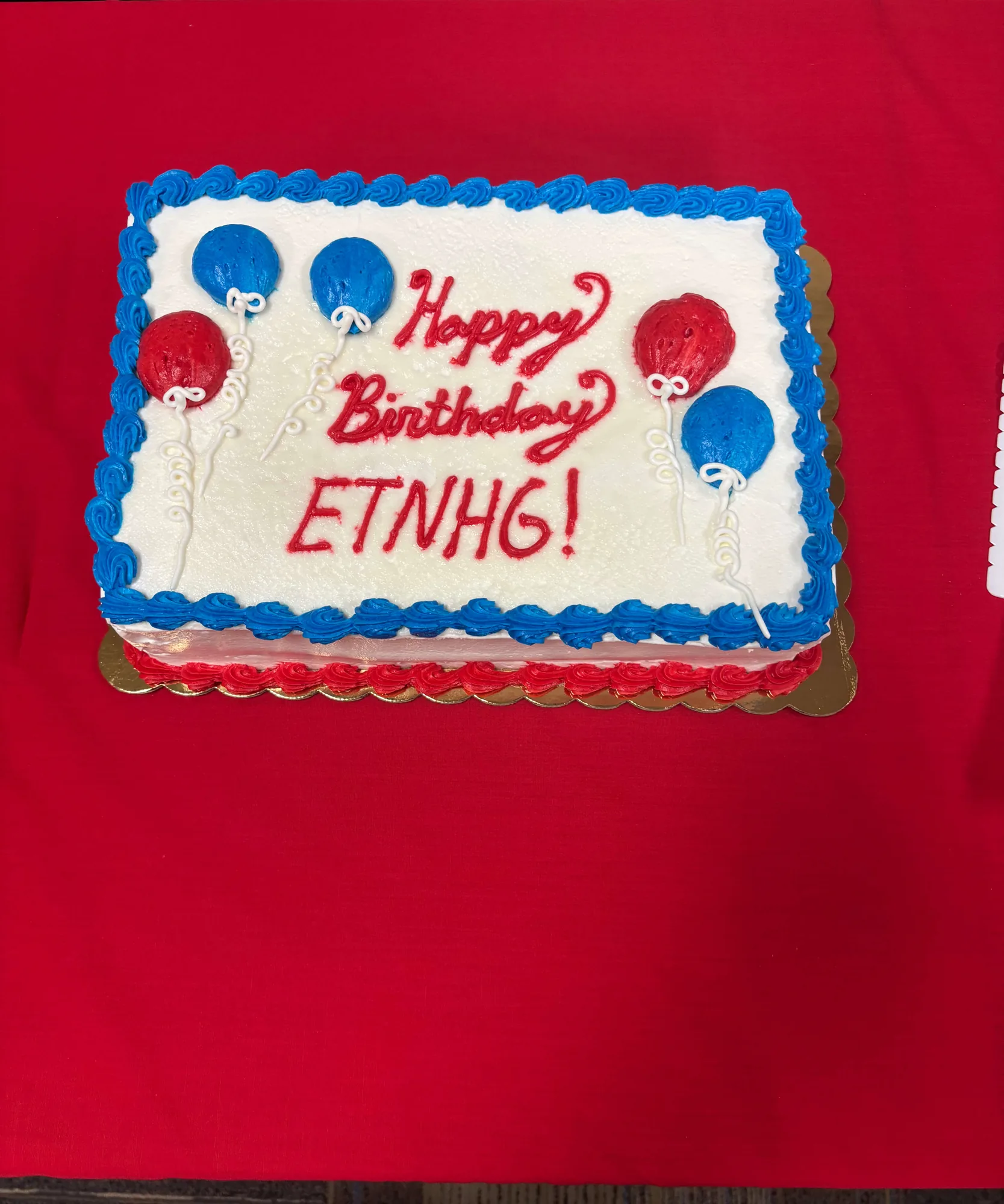 A sheet cake iced in white with a blue piped border and red and blue balloons, lettered “Happy Birthday ETNHG!” in red, on a red tablecloth.