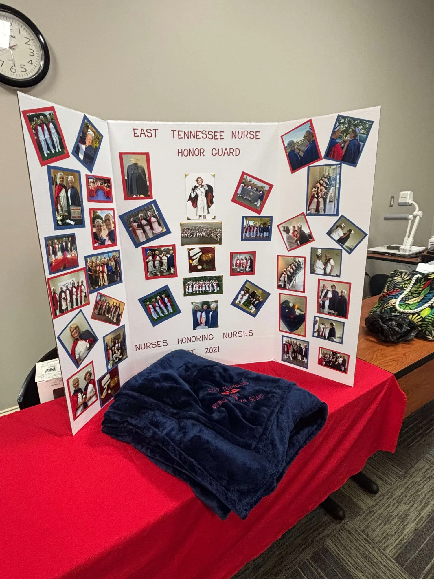 A trifold display board headed “East Tennessee Nurse Honor Guard — Nurses Honoring Nurses, Est. 2021” and covered with photographs from the chapter’s services, with a folded navy throw on the red-draped table in front of it.