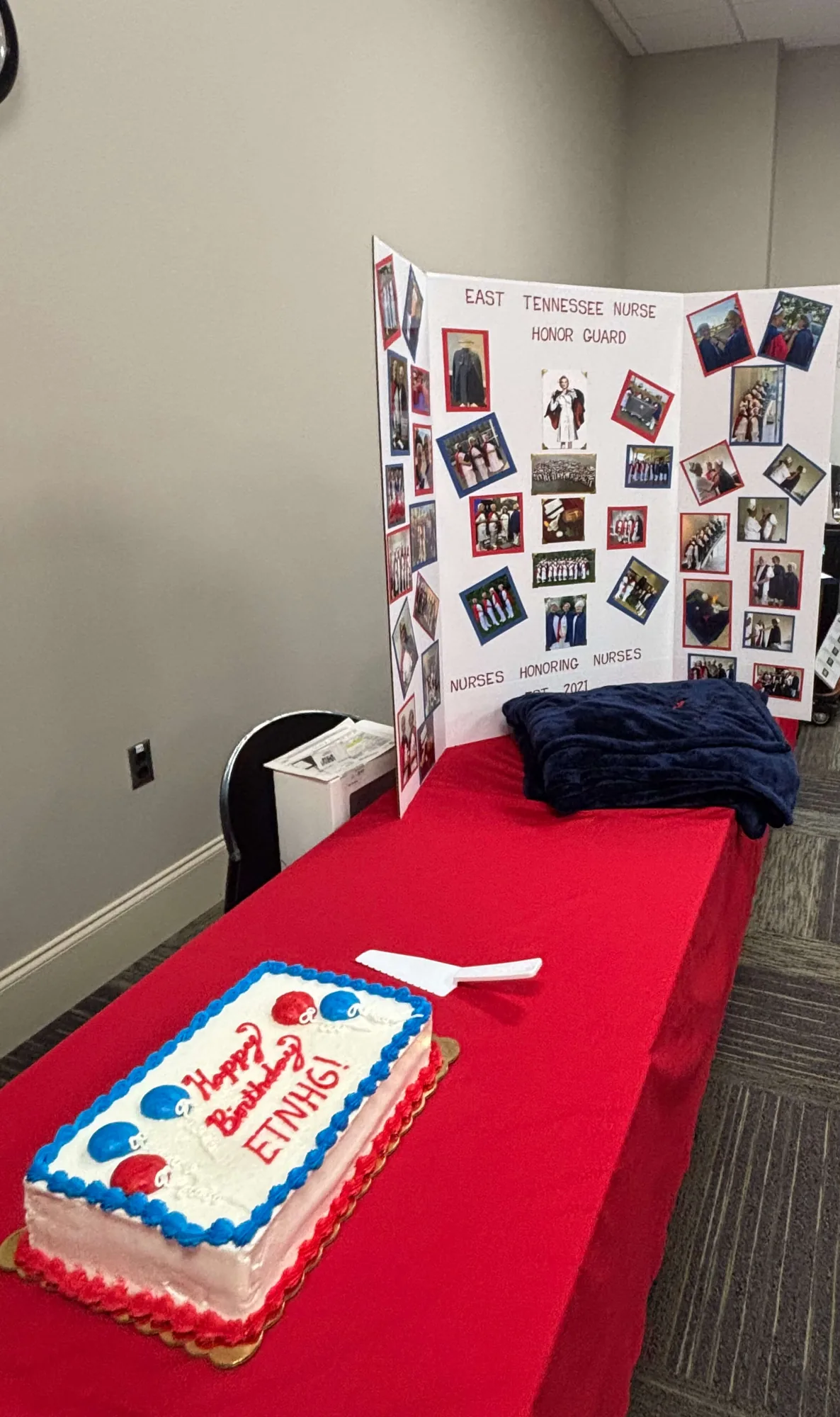 A meeting room set for the celebration: the birthday cake and a folded navy throw on a long red-draped table, with the chapter’s photograph display board standing behind.