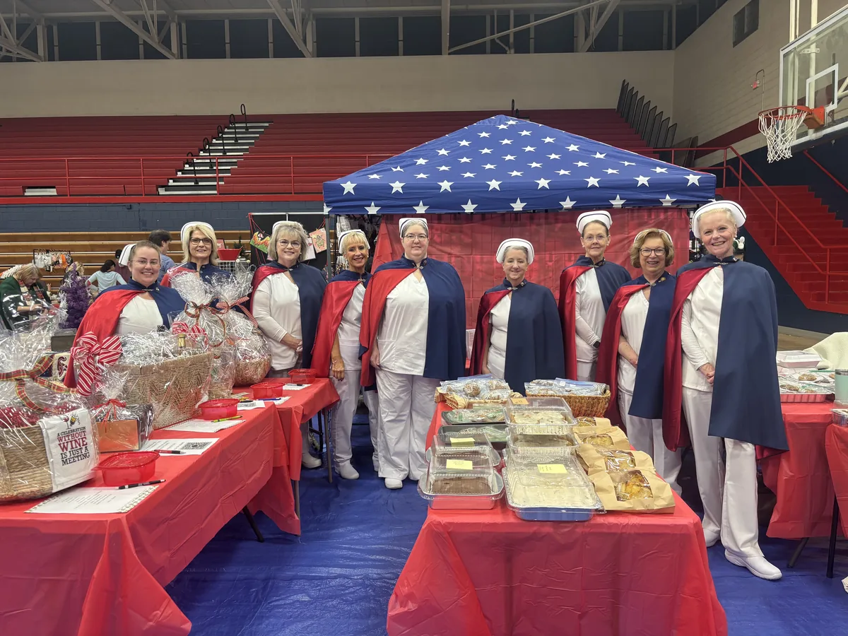 Honor guard members in uniform behind tables of baked goods under a stars and stripes canopy in a gymnasium.