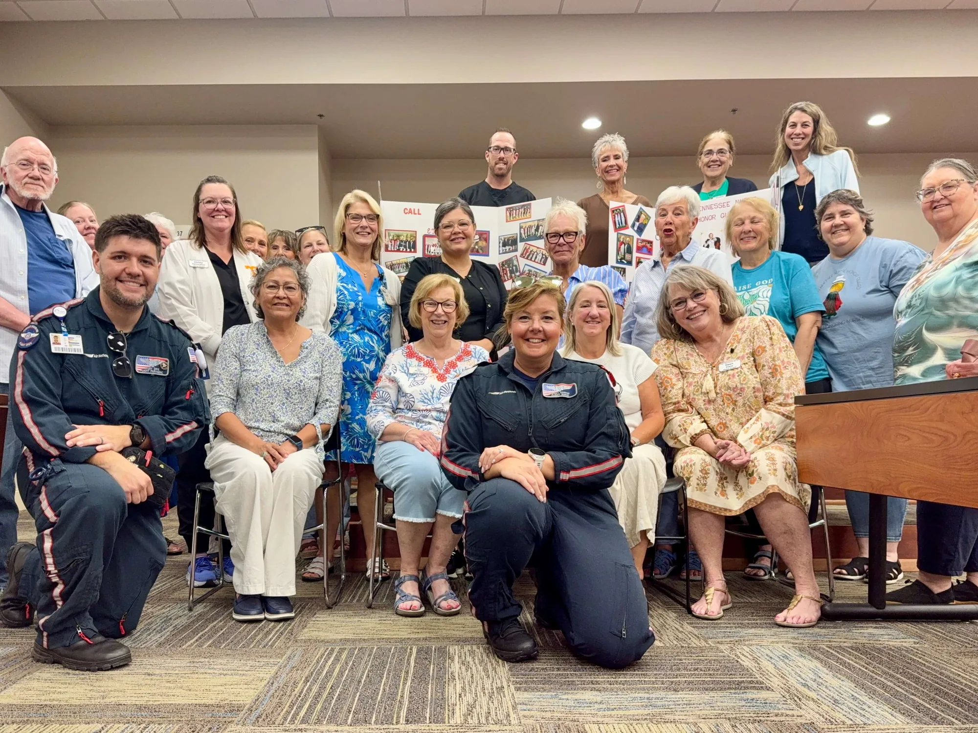 About twenty-five East Tennessee Nurse Honor Guard members gathered for a group photograph with two Covenant Health Air Medical flight crew in navy flight suits at the chapter’s August meeting, in front of the chapter’s photograph display board.