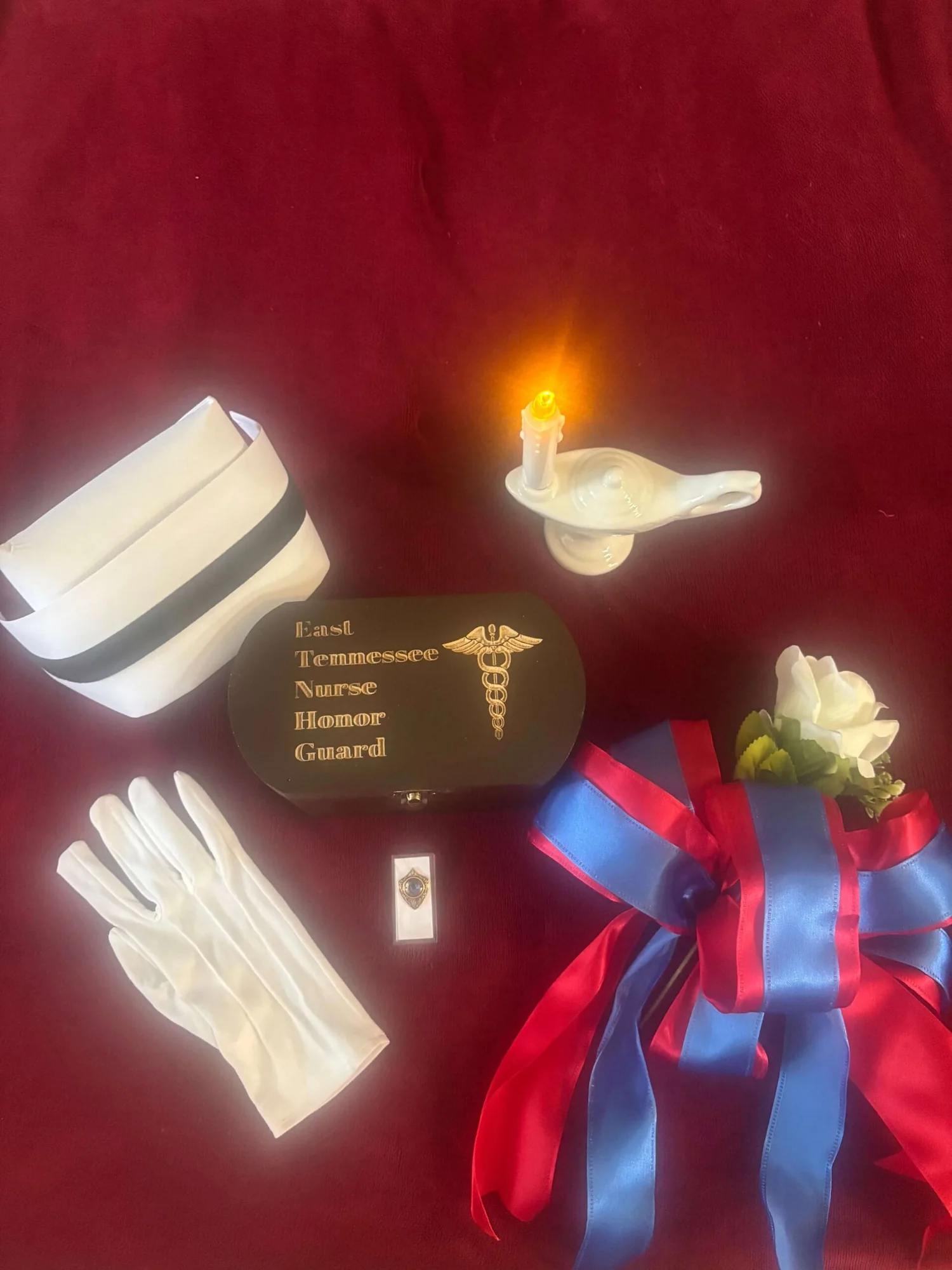 A nurse’s cap, white gloves, a Nightingale lamp with a lit candle, a service pin, a white rose and a red-and-blue ribbon arranged on red velvet around an engraved East Tennessee Nurse Honor Guard box.