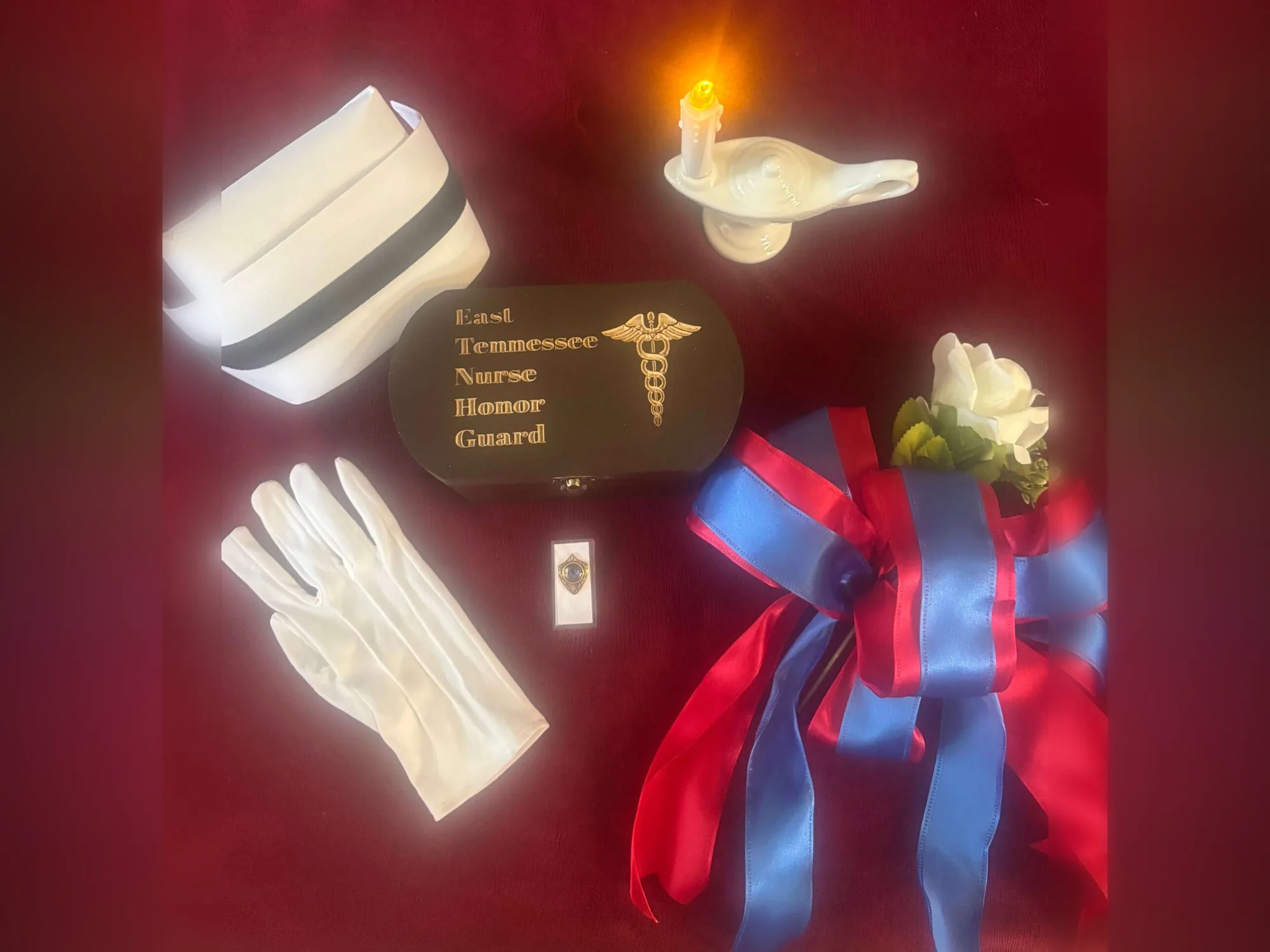 A nurse’s cap, white gloves, a Nightingale lamp with a lit candle, a service pin, a white rose and a red-and-blue ribbon arranged on red velvet around an engraved East Tennessee Nurse Honor Guard box.