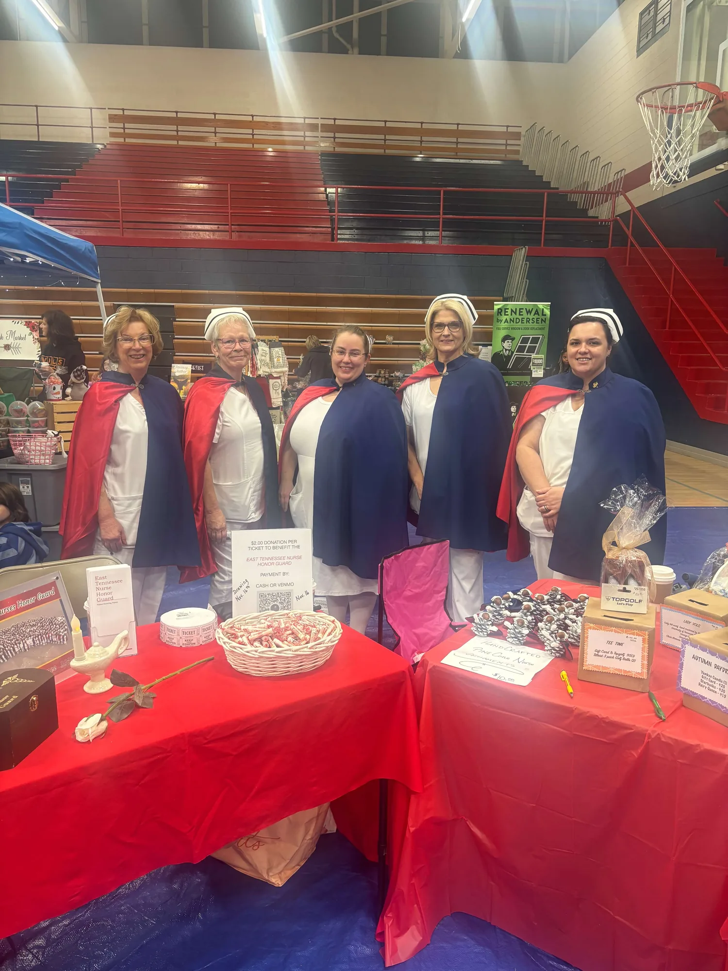 Five honor guard members in capes standing behind a red-draped raffle table of gift baskets in a school gymnasium in 2024.