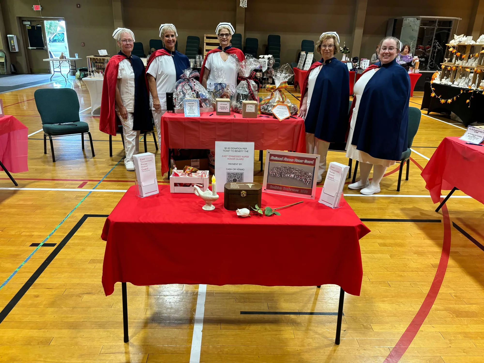 A red-draped fundraiser table holding a donation box and framed information about the chapter, with honor guard members standing behind it in a gymnasium.