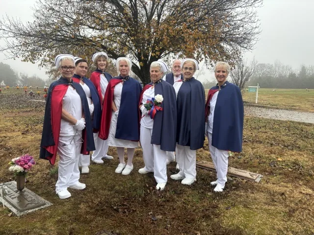 Nine honor guard members in capes standing at a graveside beneath a bare tree on a foggy morning, one of them holding a white rose tied with red, white and blue ribbon.