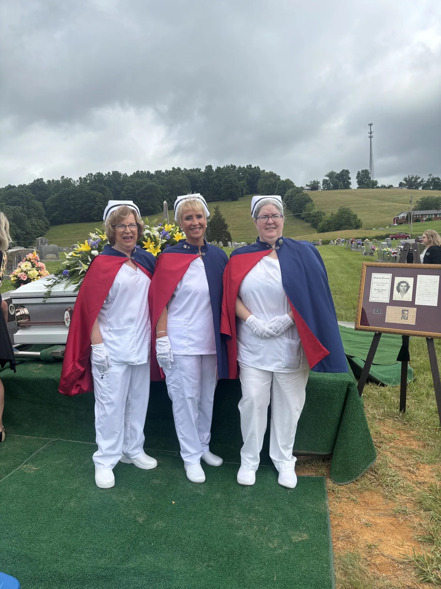 Three honor guard members in white uniforms, capes and white gloves standing beside the flowers at a graveside service on a hillside.
