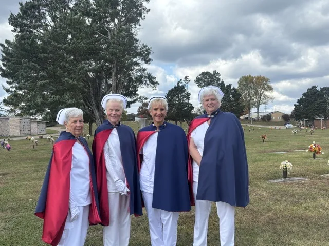 Four honor guard members in white uniforms and red-and-navy capes standing on the lawn of a memorial park under a cloudy sky.