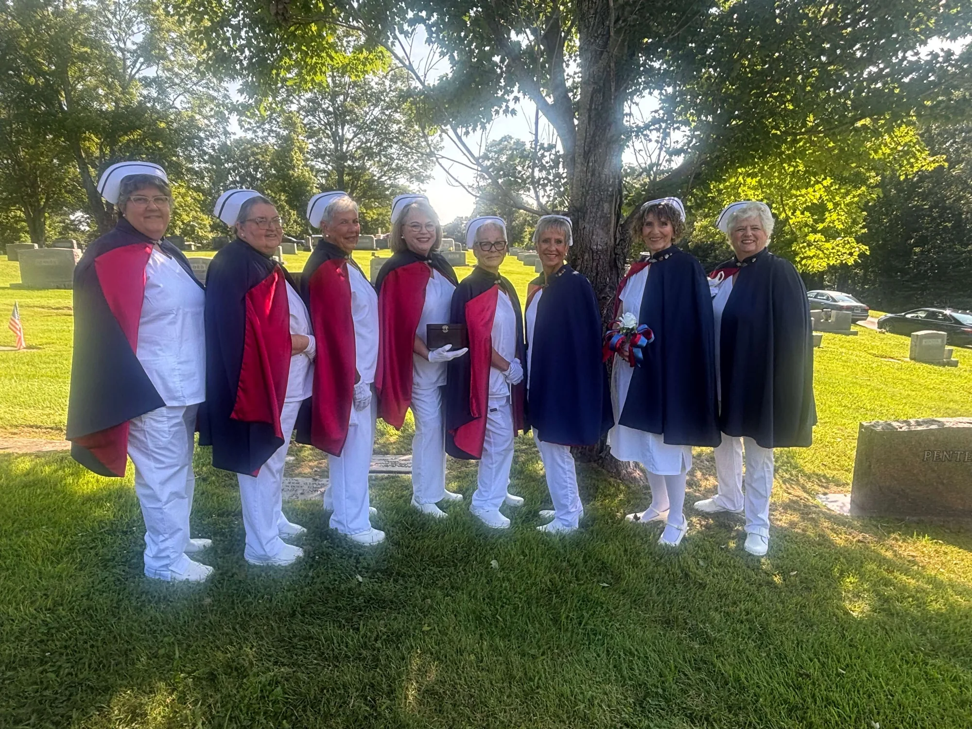 Eight honor guard members standing in a line on the grass beneath trees at a graveside tribute.