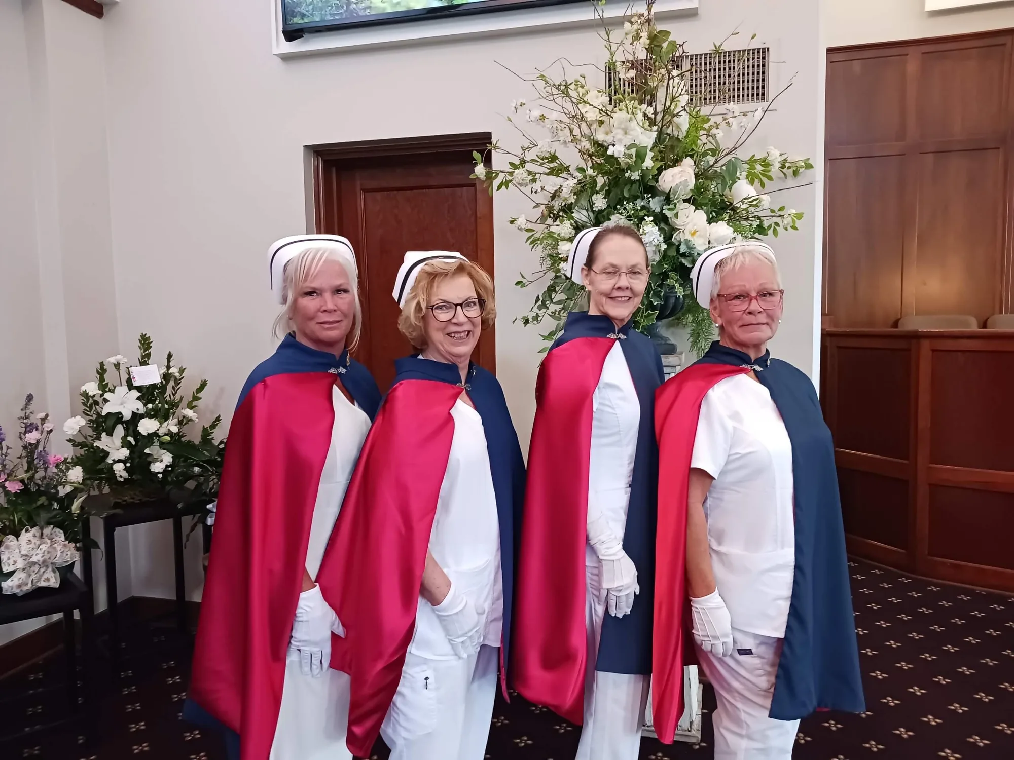 Four honor guard members in white uniforms and capes standing beside a tall white floral arrangement at a service.