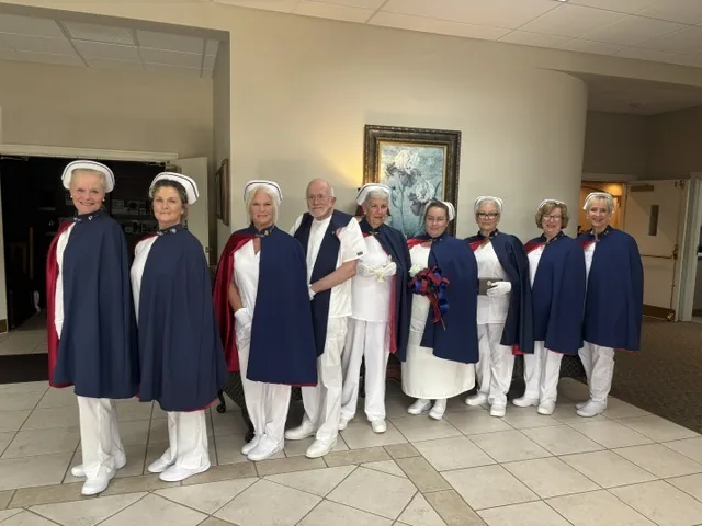 Nine honor guard members standing in line in a funeral home lobby, one of them in a white dress uniform holding a white rose tied with ribbon.