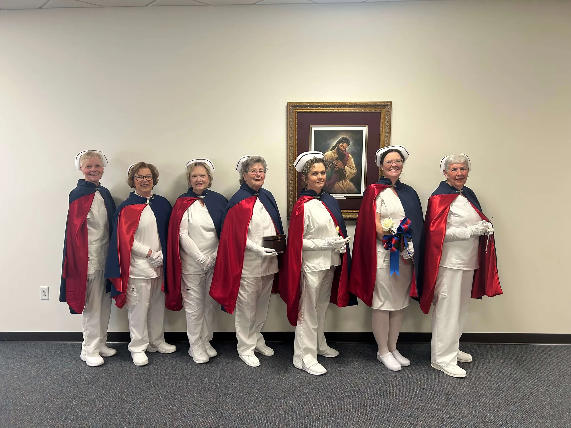 Seven honor guard members standing in a line in white uniforms and capes beneath a framed painting at a memorial service.