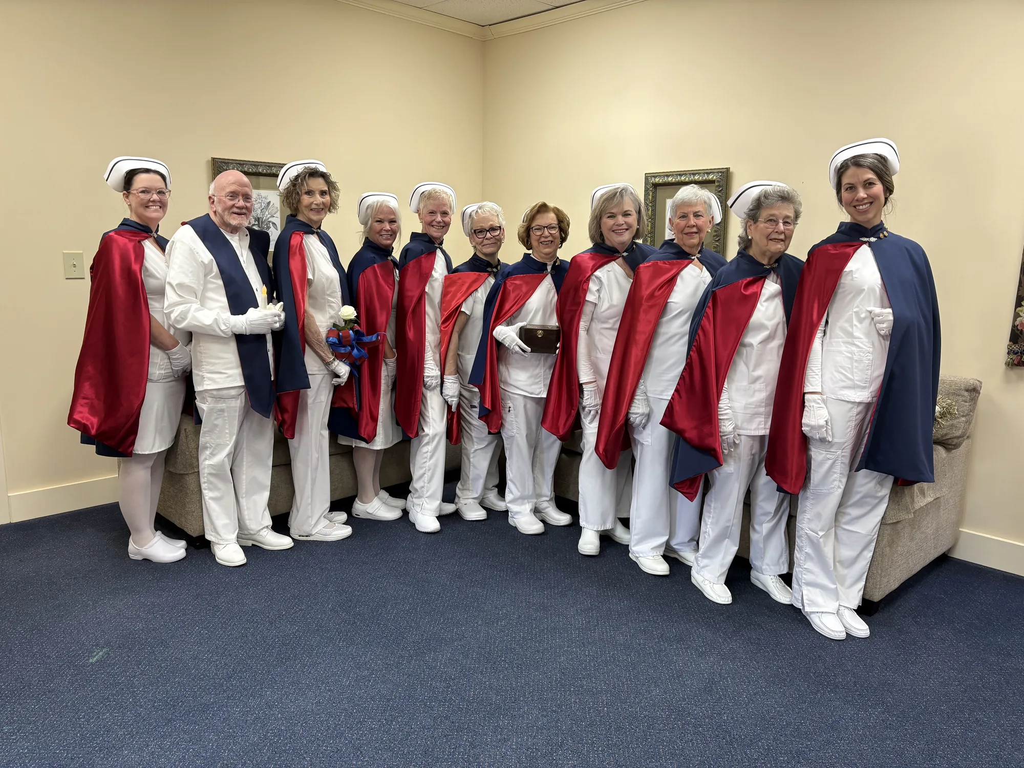 Ten honor guard members standing shoulder to shoulder in white uniforms and red and navy capes in a funeral home chapel.