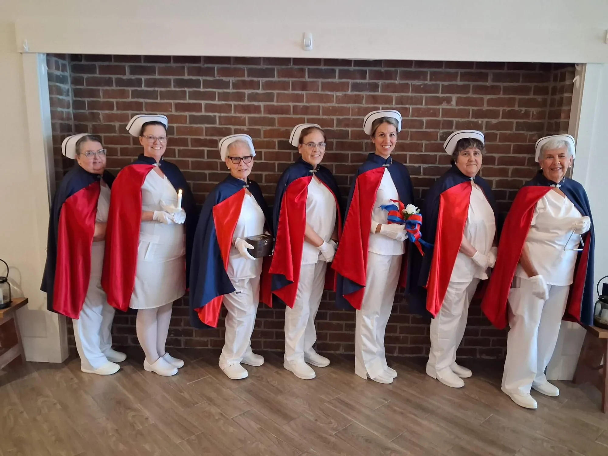 Seven honor guard members standing shoulder to shoulder in white uniforms and red and navy capes against a brick wall.