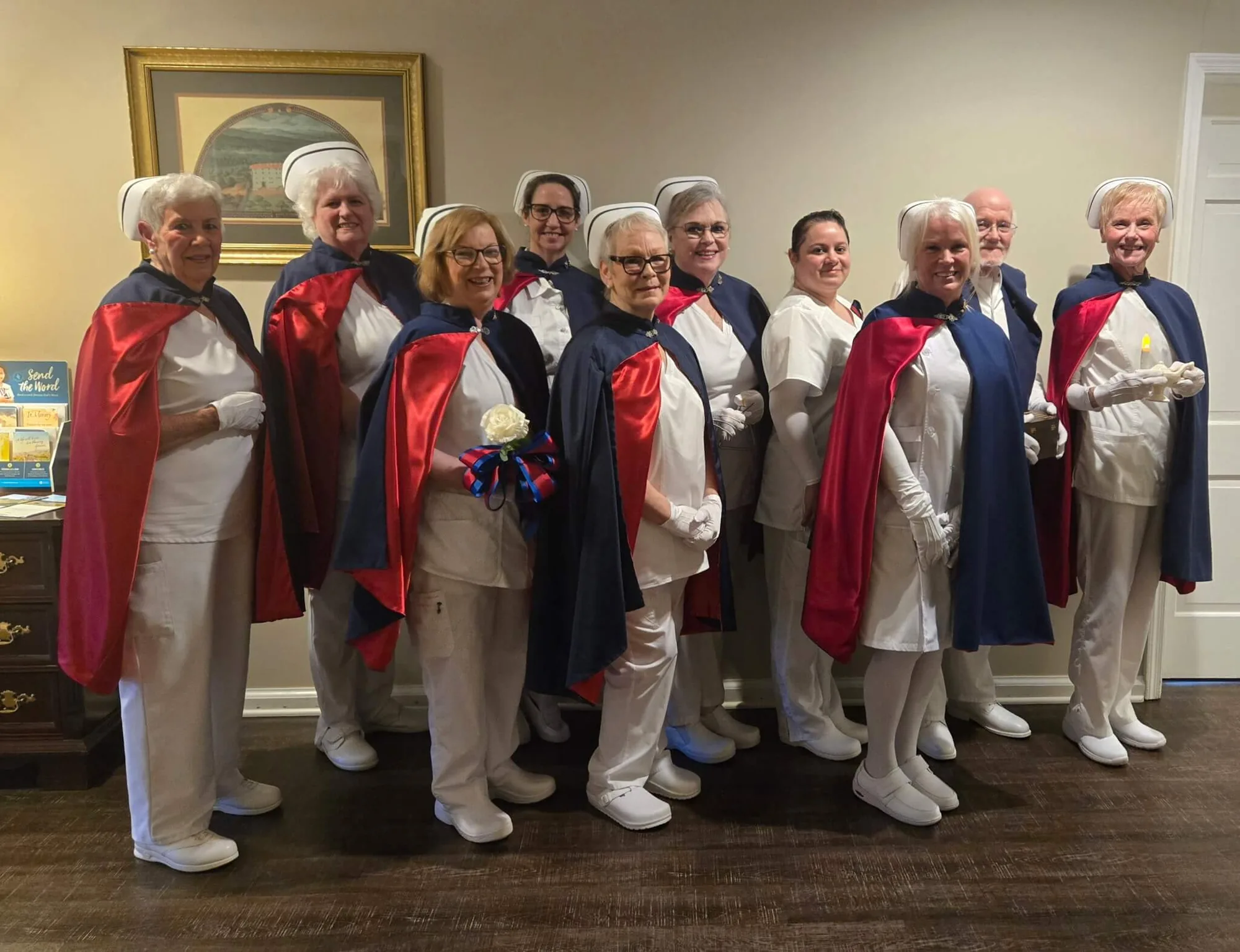 Nine honor guard members standing in a line in white uniforms and red and navy capes in a funeral home lobby.