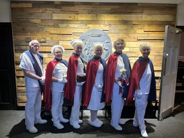 Six honor guard members standing in line before a reclaimed-wood wall in a funeral home, one holding the Nightingale lamp and another a white rose.