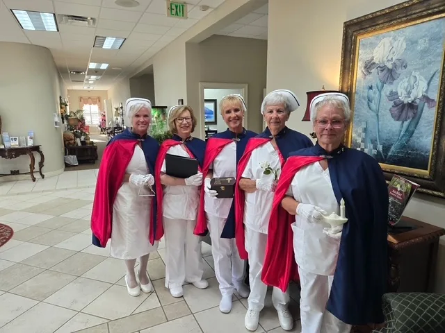 Five honor guard members in a funeral home lobby, one holding the Nightingale lamp, another a service folder and a third a white rose.