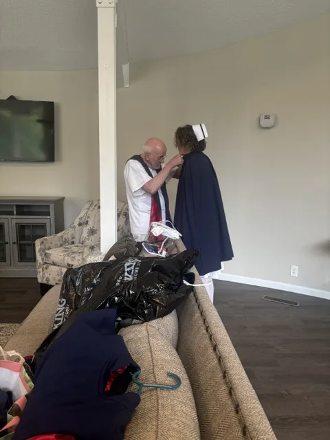 A member of the honor guard fastening the collar of another member's navy cape as the guard dresses for a service, garment bags laid over the sofa behind them.