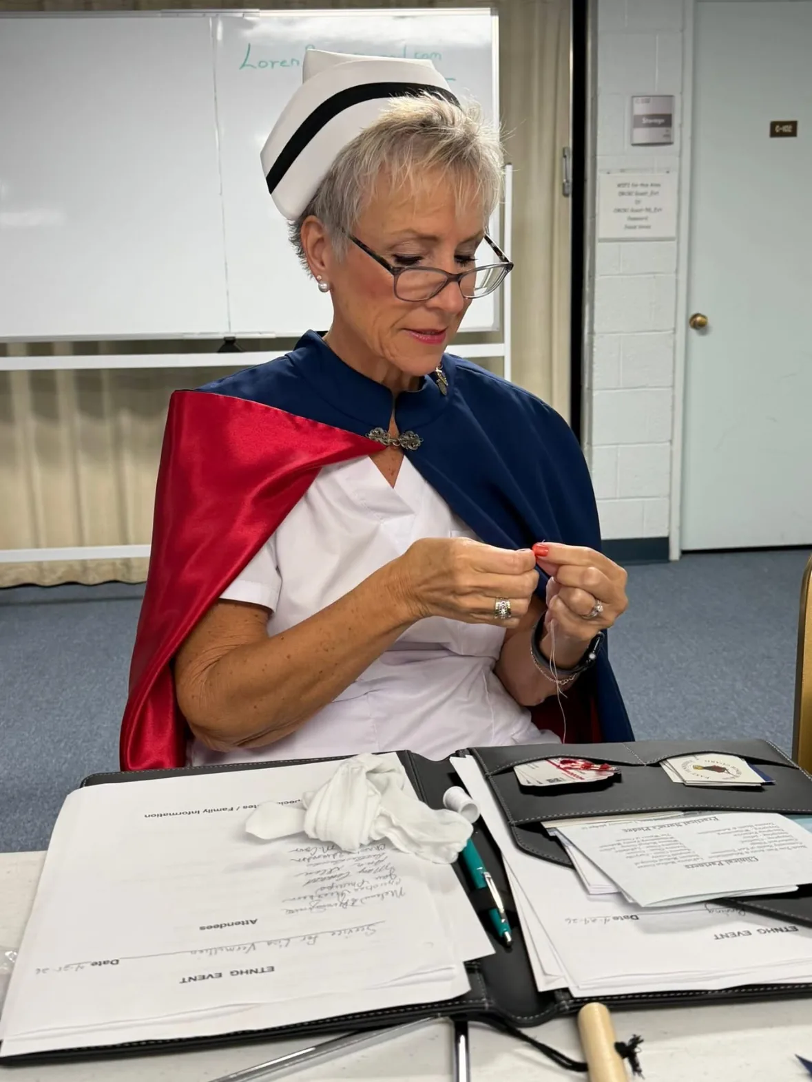 A member of the East Tennessee Nurse Honor Guard, in white uniform and navy cape, sitting at a table threading a needle to mend a white glove before a ceremony.