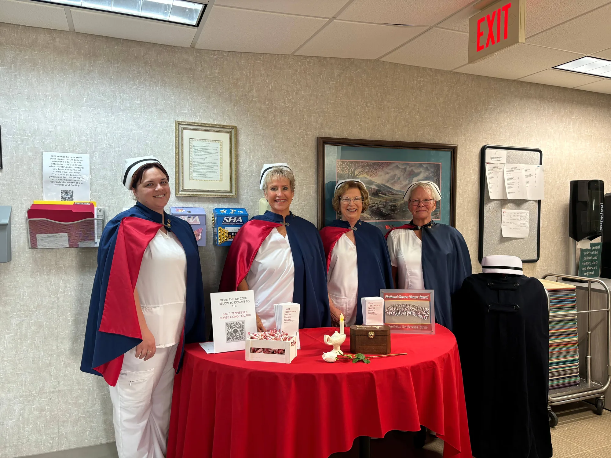 Four honor guard members in uniform standing beside a red-draped information table in a hospital lobby in 2024.