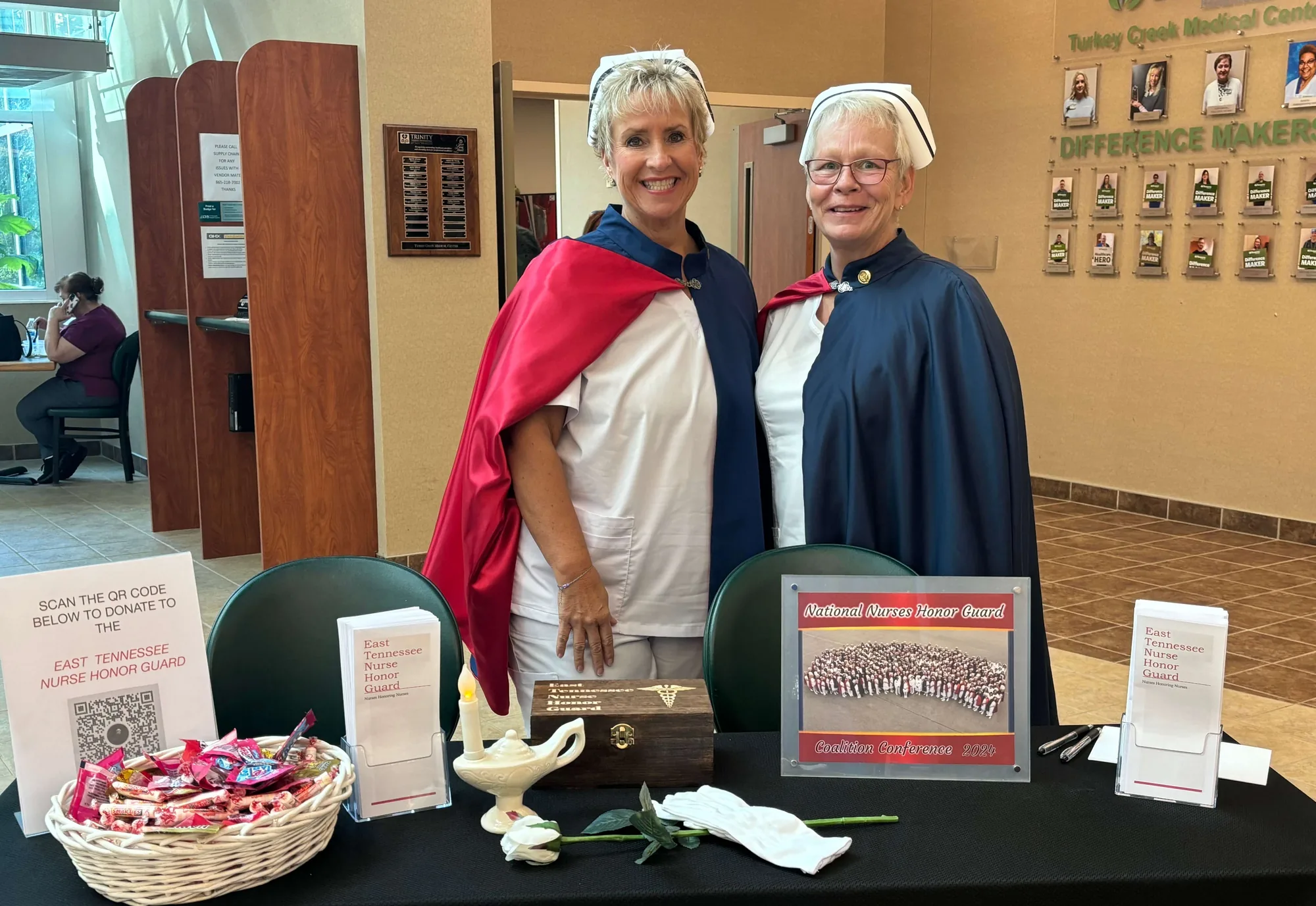 Two honor guard members in capes behind an information table set with a white rose, a Nightingale lamp and printed information about the chapter, in a hospital lobby.