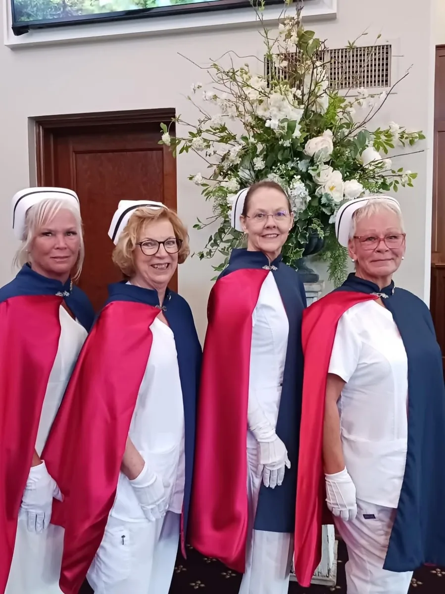 Four East Tennessee Nurse Honor Guard members in white uniforms, capes and white gloves standing beside a large white floral arrangement at a service.