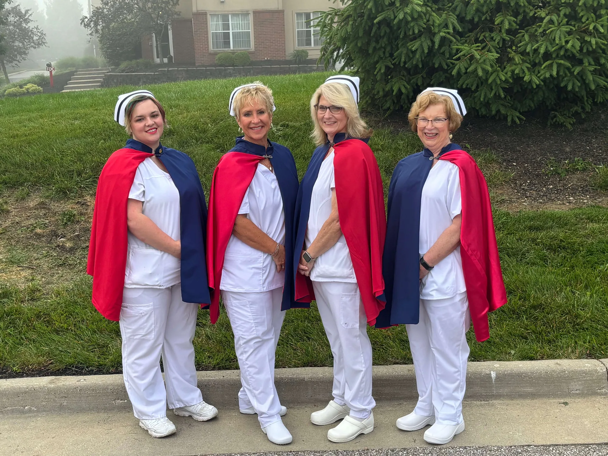 Four East Tennessee Nurse Honor Guard members in white uniforms and red and navy capes standing together on a misty lawn at the 2024 national convention.