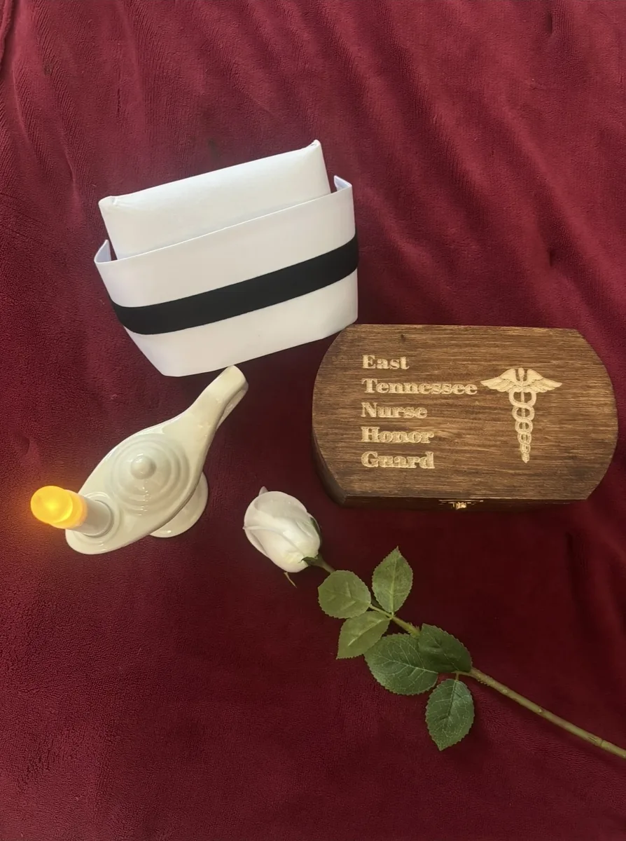 A nurse's cap, a single white rose, and a Nightingale lamp arranged on red fabric beside an engraved East Tennessee Nurse Honor Guard plaque.