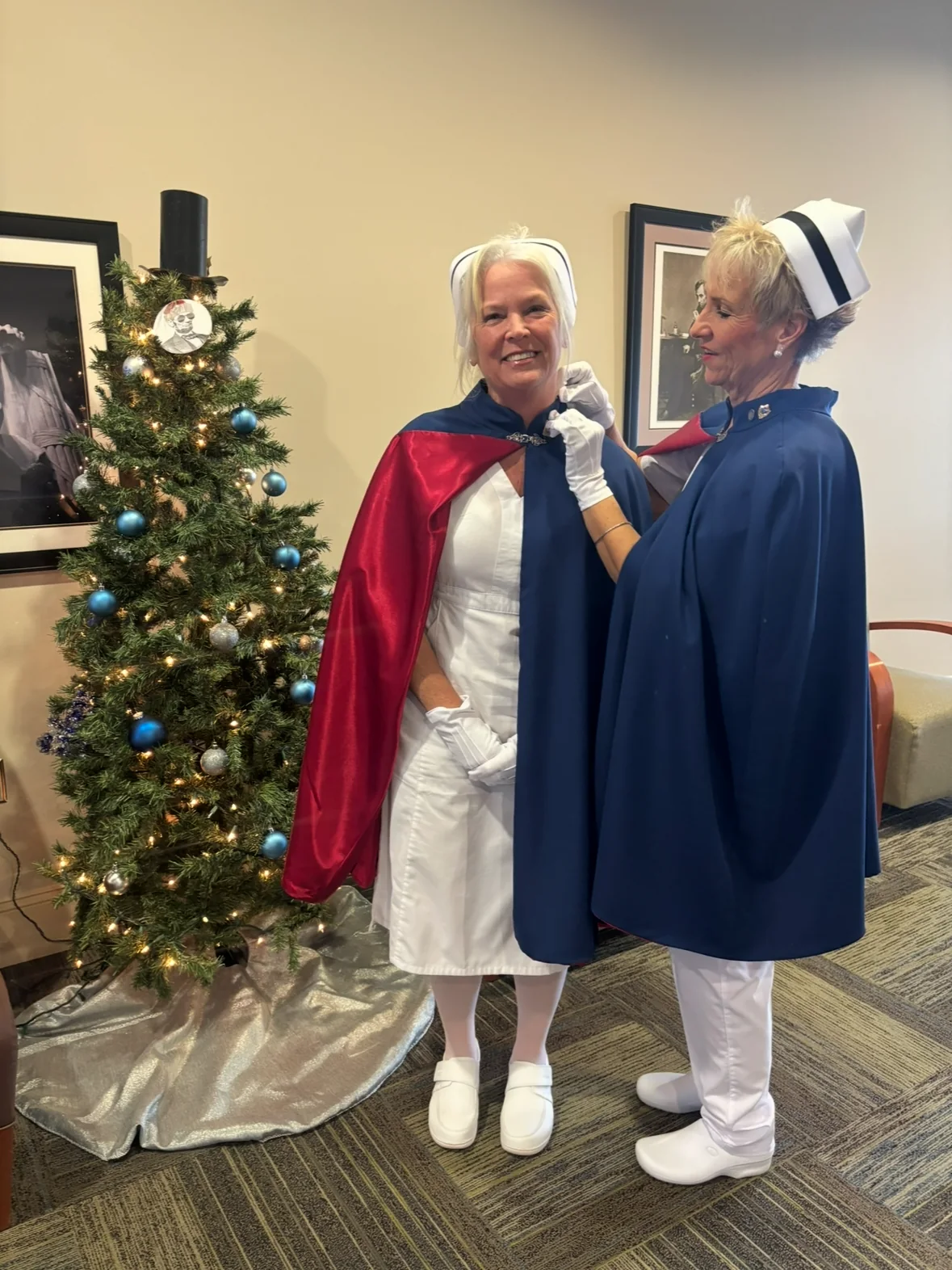 Cathy Felker in a white nursing uniform and red-lined cape receiving her first-service pin from Melanie Pfennigwerth beside a decorated Christmas tree.