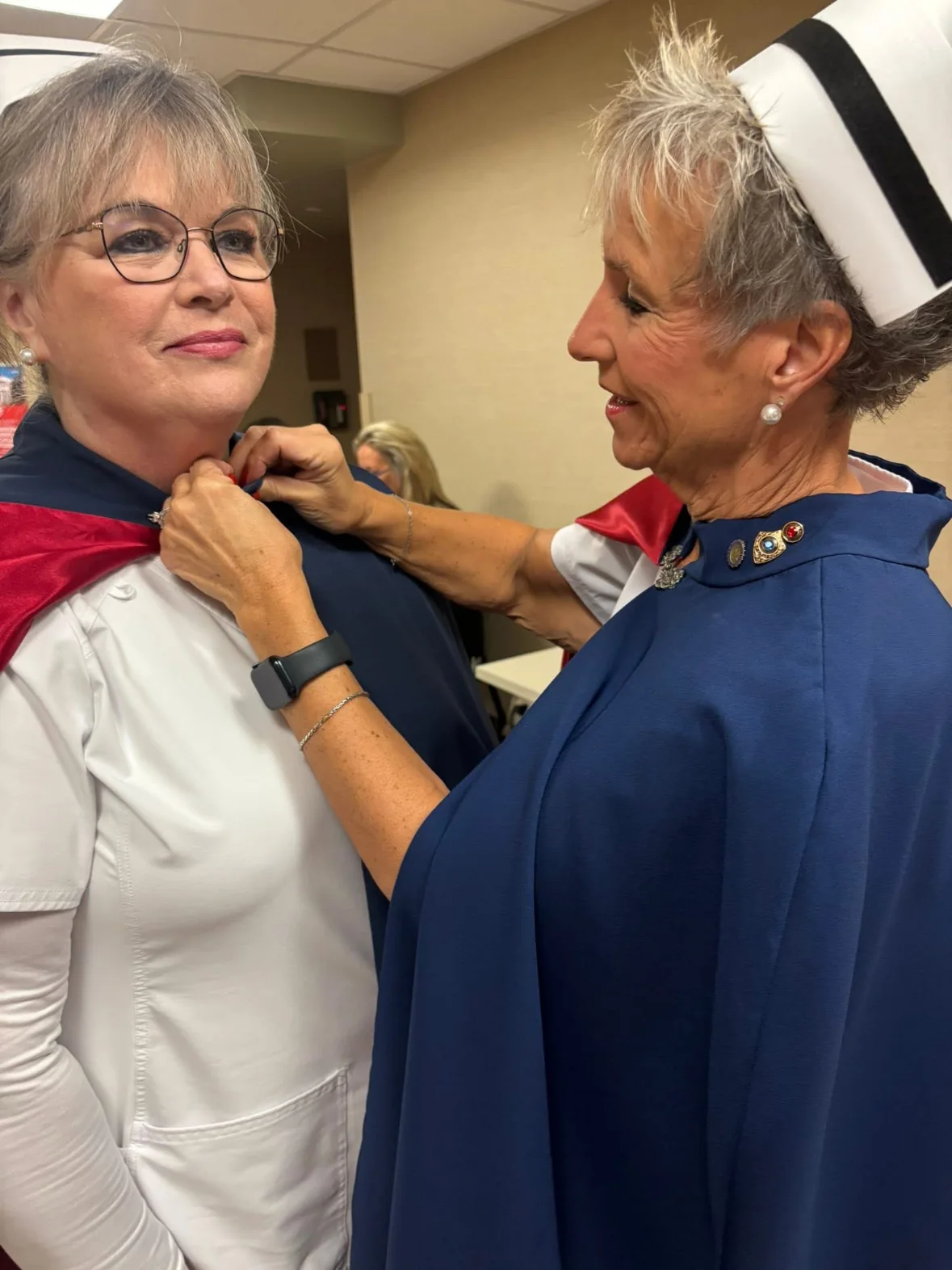 Dawn Evans in a white uniform and cape smiling as Melanie Pfennigwerth fastens her 25-service pin at her collar.