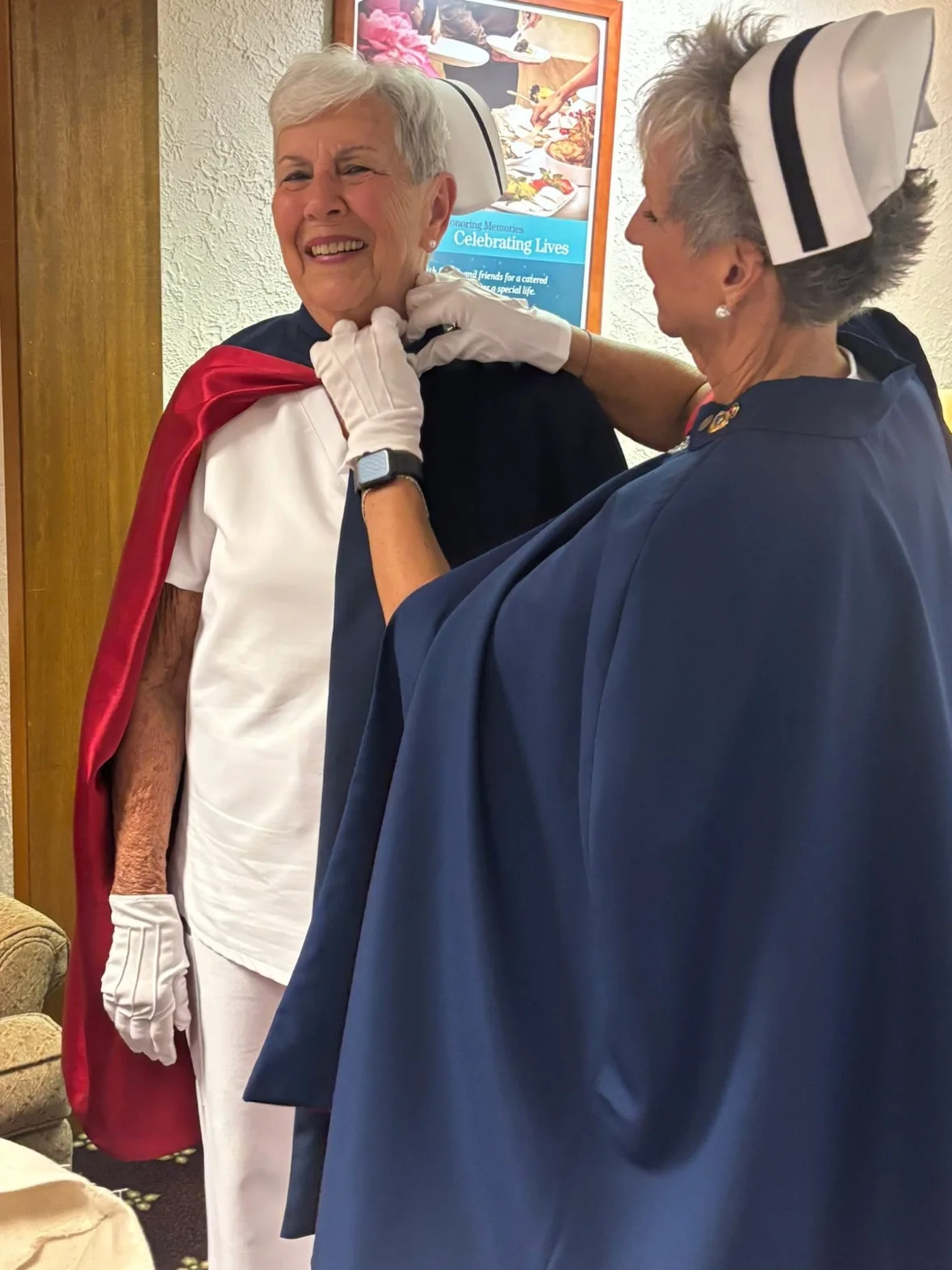 Ann Allen in a white uniform and red-lined cape smiling as Melanie Pfennigwerth pins her 50-service pin.