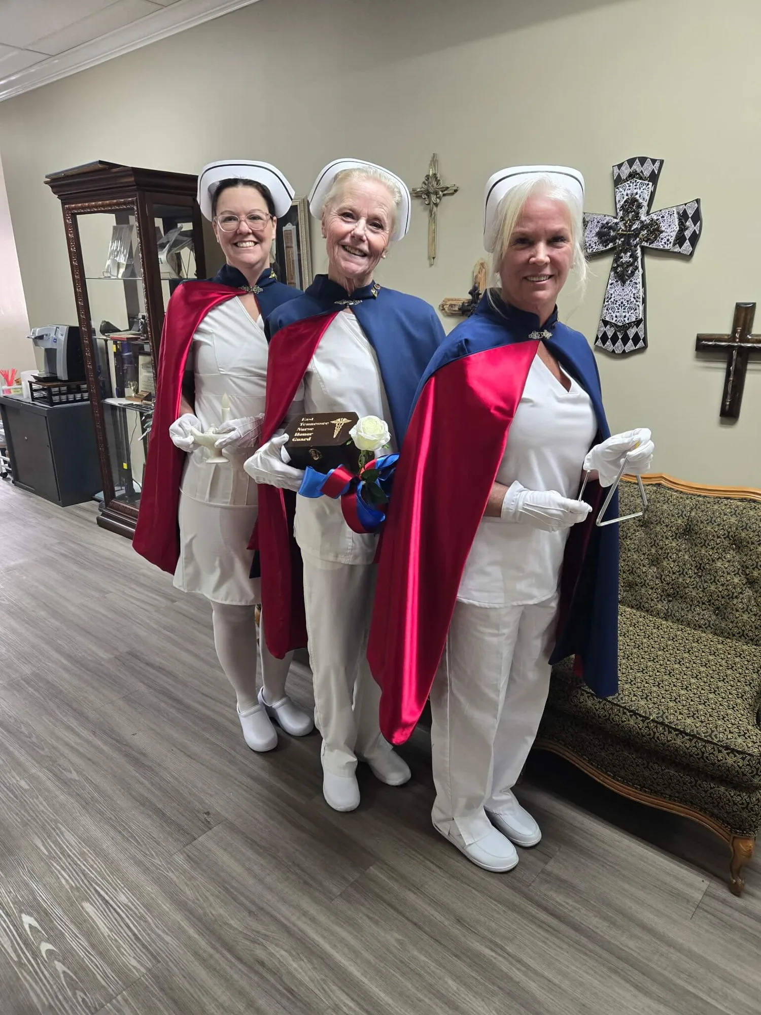 Three honor guard members in white uniforms and capes, one holding the Nightingale lamp, standing in a funeral home foyer.
