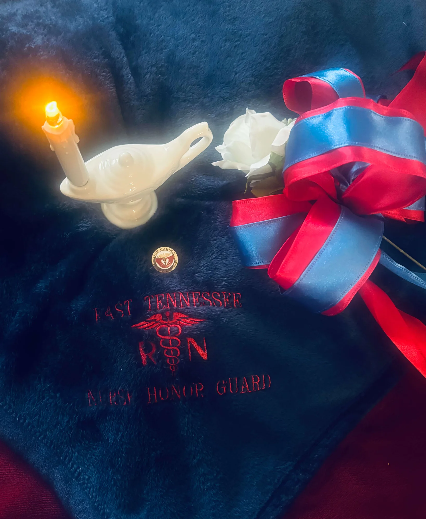 A Nightingale lamp with a lit candle, a white rose, a red-and-blue ribbon rosette and a round pin reading “Our Caring Makes a Difference”, arranged on the navy East Tennessee Nurse Honor Guard throw.