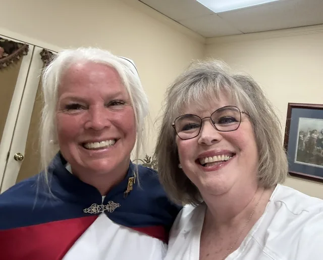 Two honor guard members leaning together for a photograph, one of them wearing a red-and-navy cape.