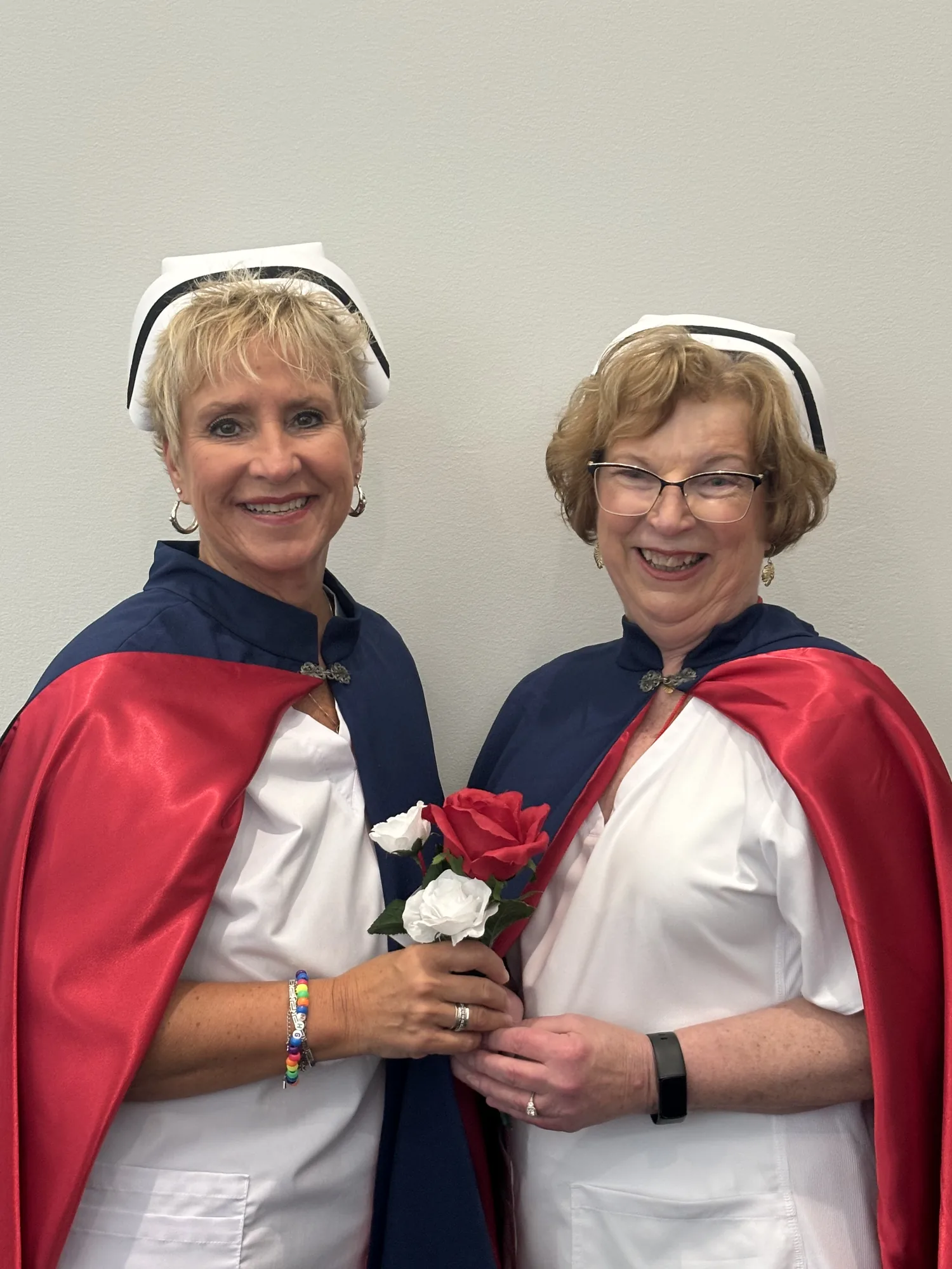Two East Tennessee Nurse Honor Guard members in white uniforms and red and navy capes standing together, holding a red rose and a white rose.