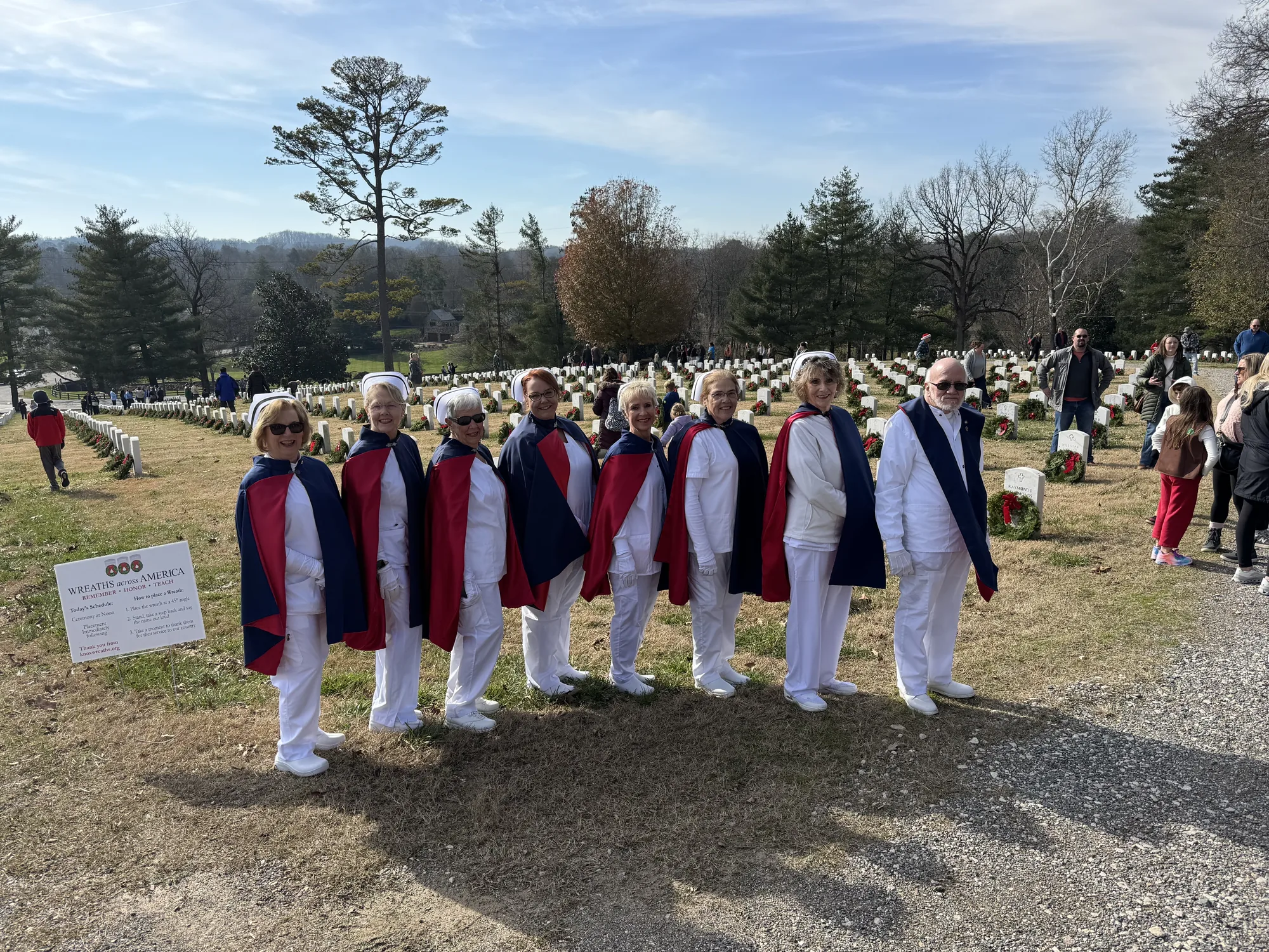 East Tennessee Nurse Honor Guard members in uniform and capes standing in a line beside rows of white headstones, each laid with an evergreen remembrance wreath, at a national cemetery on a bright winter day.
