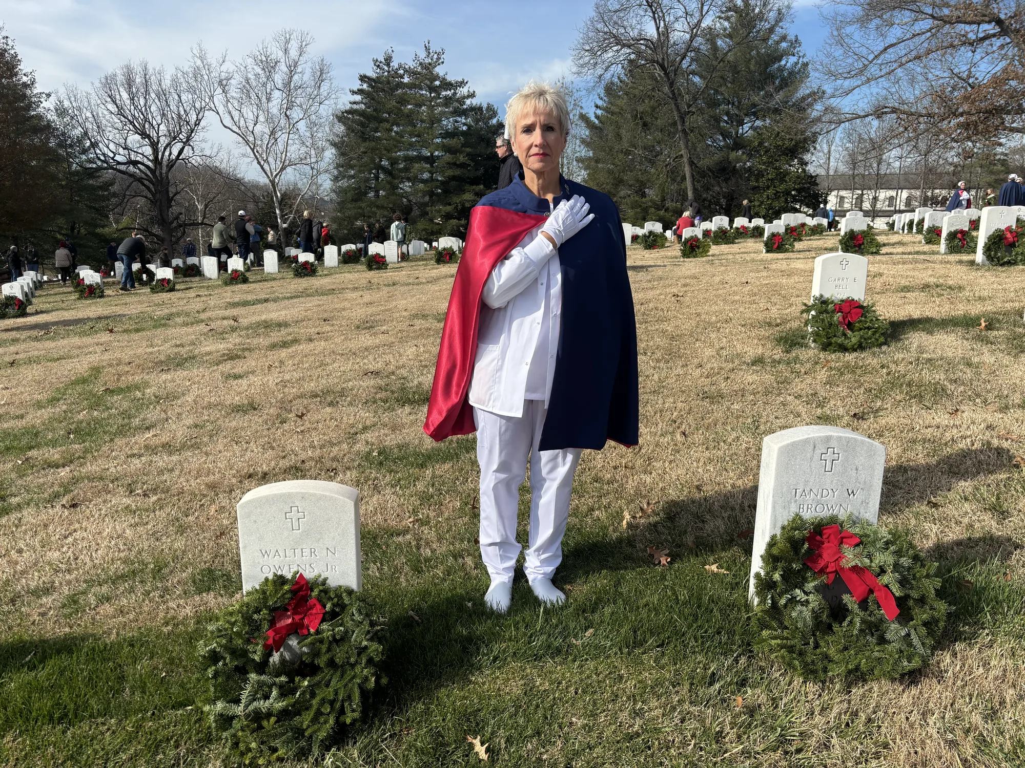 An East Tennessee Nurse Honor Guard member in white uniform and cape standing with a hand over the heart between two wreath-laid headstones at a national cemetery.