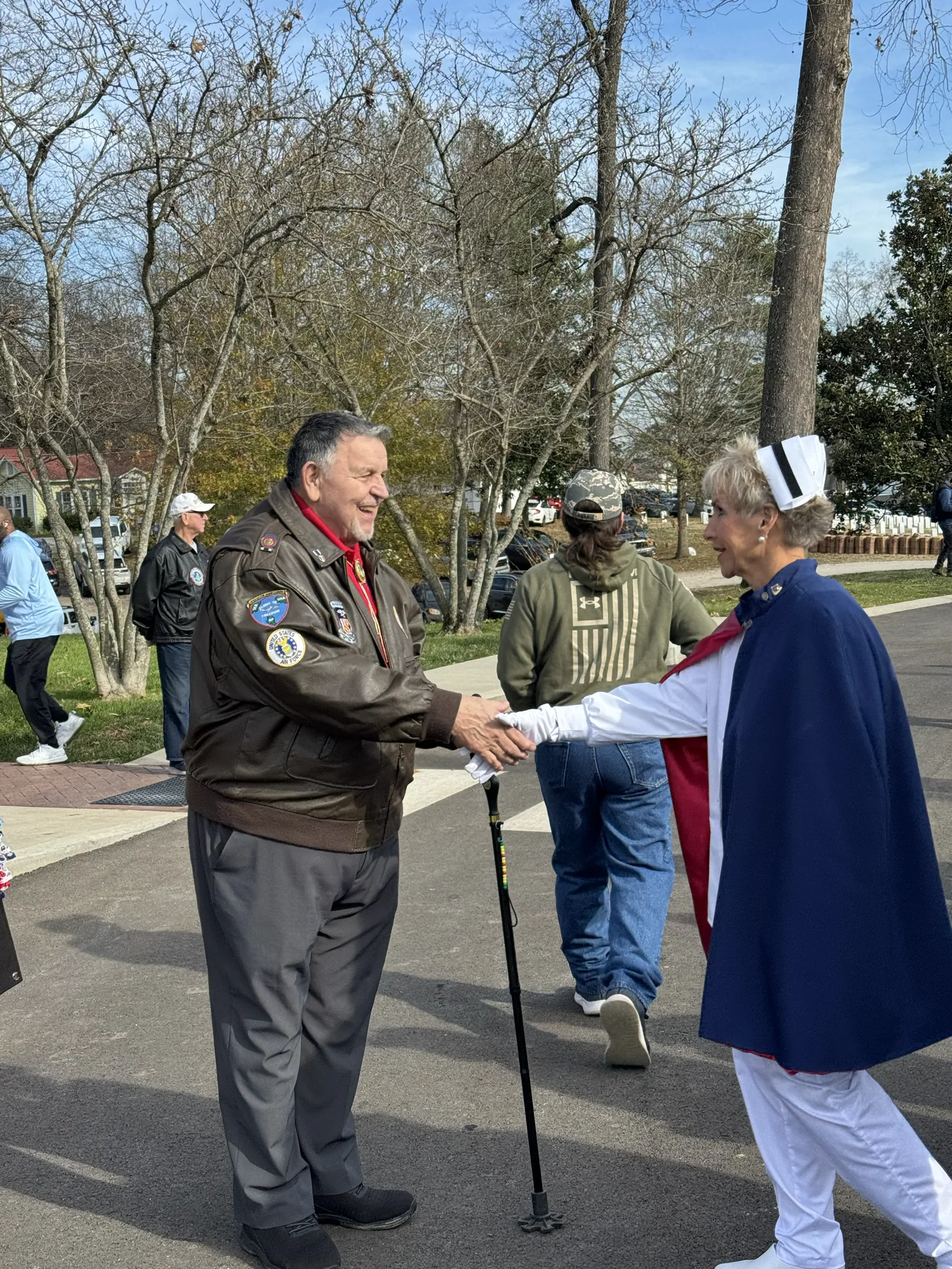 An honor guard member in uniform and cape shaking the hand of a veteran in a patched flight jacket at a Wreaths Across America ceremony.