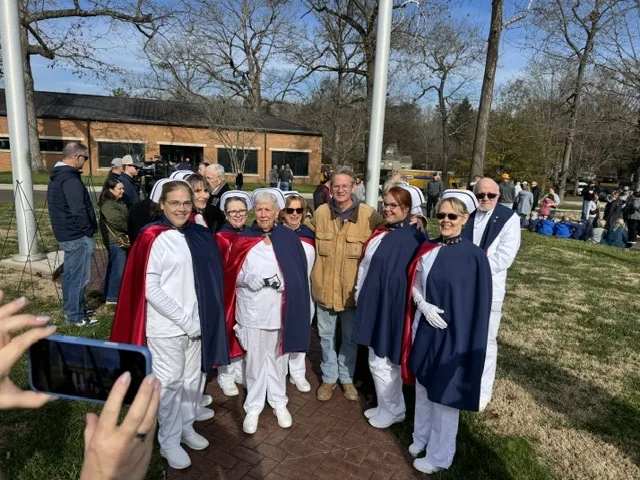 Honor guard members gathered outdoors with a guest at a Wreaths Across America ceremony while another member photographs them.