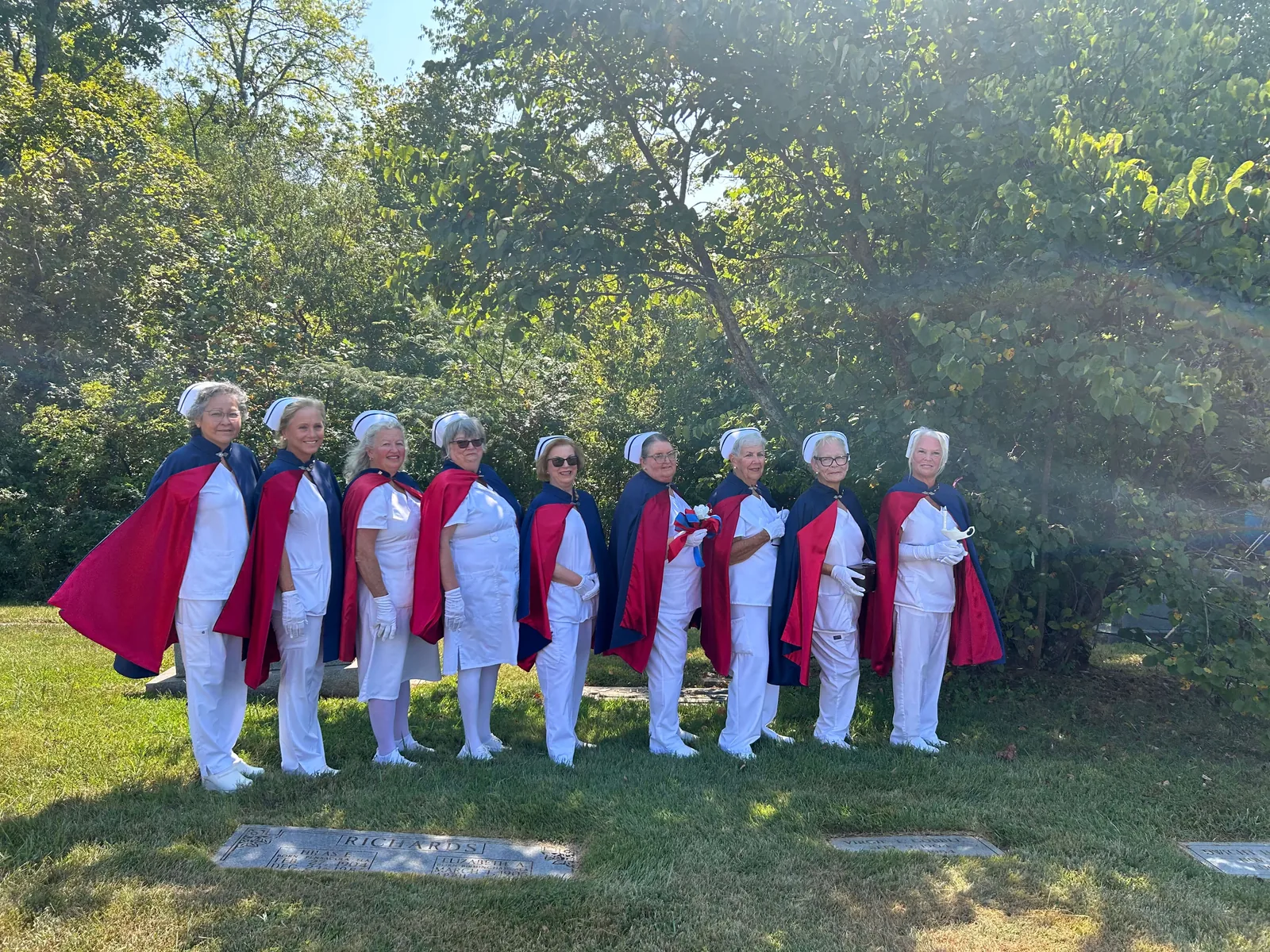 Nine honor guard members in white uniforms, white nurses’ caps and navy capes lined in red, standing in a row at a graveside beneath trees on a sunny day. One holds a white rose tied with red, white and blue ribbon, one the dark wooden presentation box, and one the white Nightingale lamp.