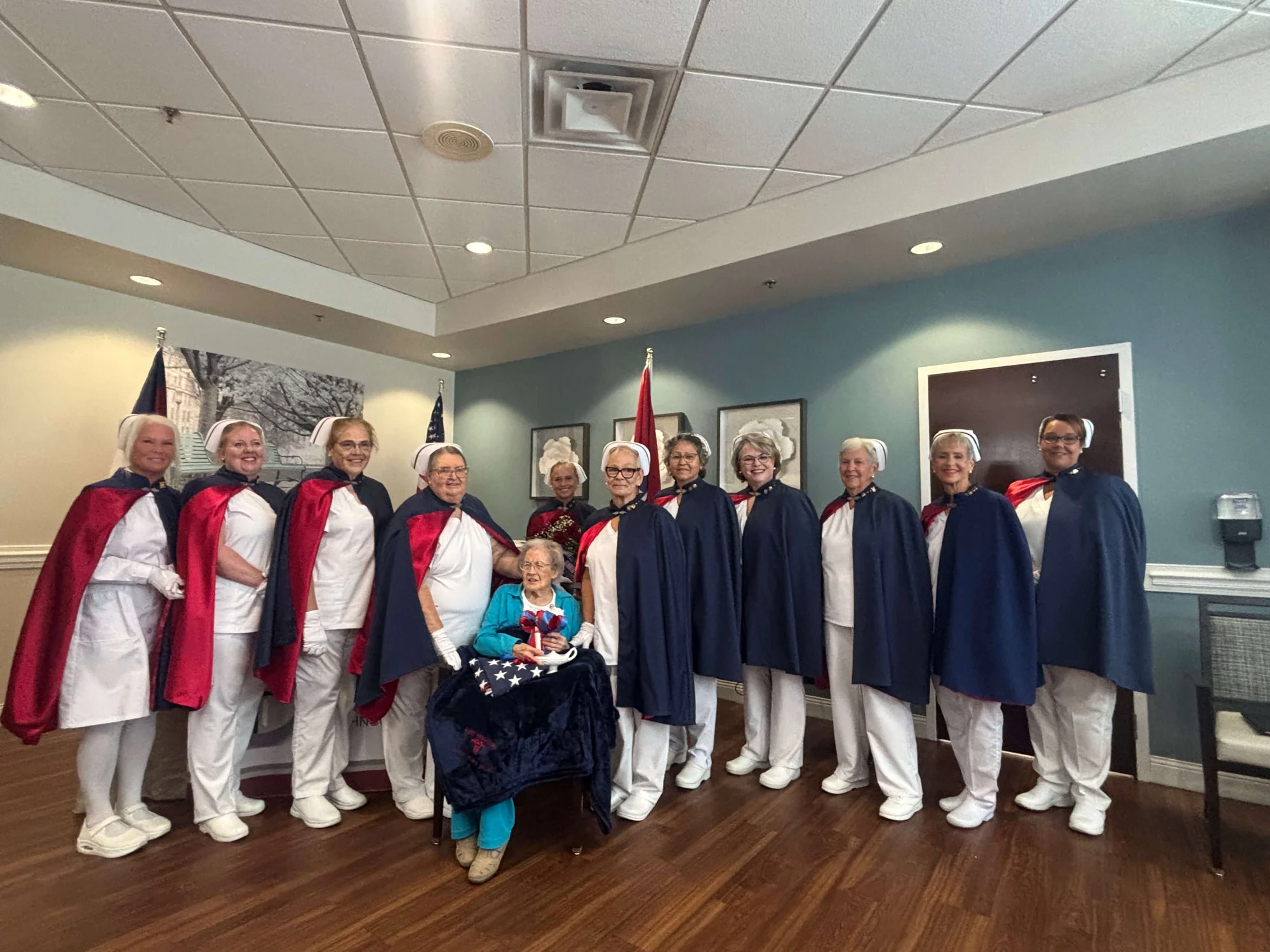 Agnes Lowe, seated in a teal outfit with the embroidered navy Honor Guard blanket across her lap, holding a folded American flag, a Nightingale lamp and a ribboned rose, surrounded by eleven East Tennessee Nurse Honor Guard members in white uniforms, white caps and navy-and-red capes, at her Living Tribute on 10 September 2026.
