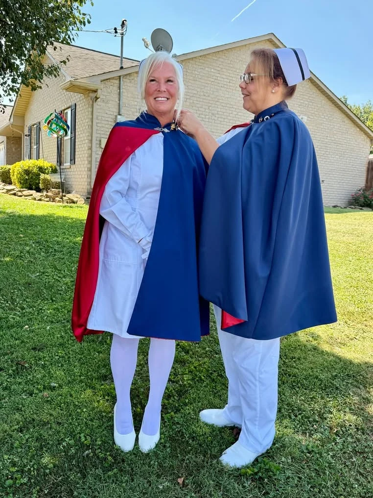 Cathy Felker in a white uniform and red-lined cape smiling outdoors as Christina Trentham, in cape and banded cap, pins her for 50 services.