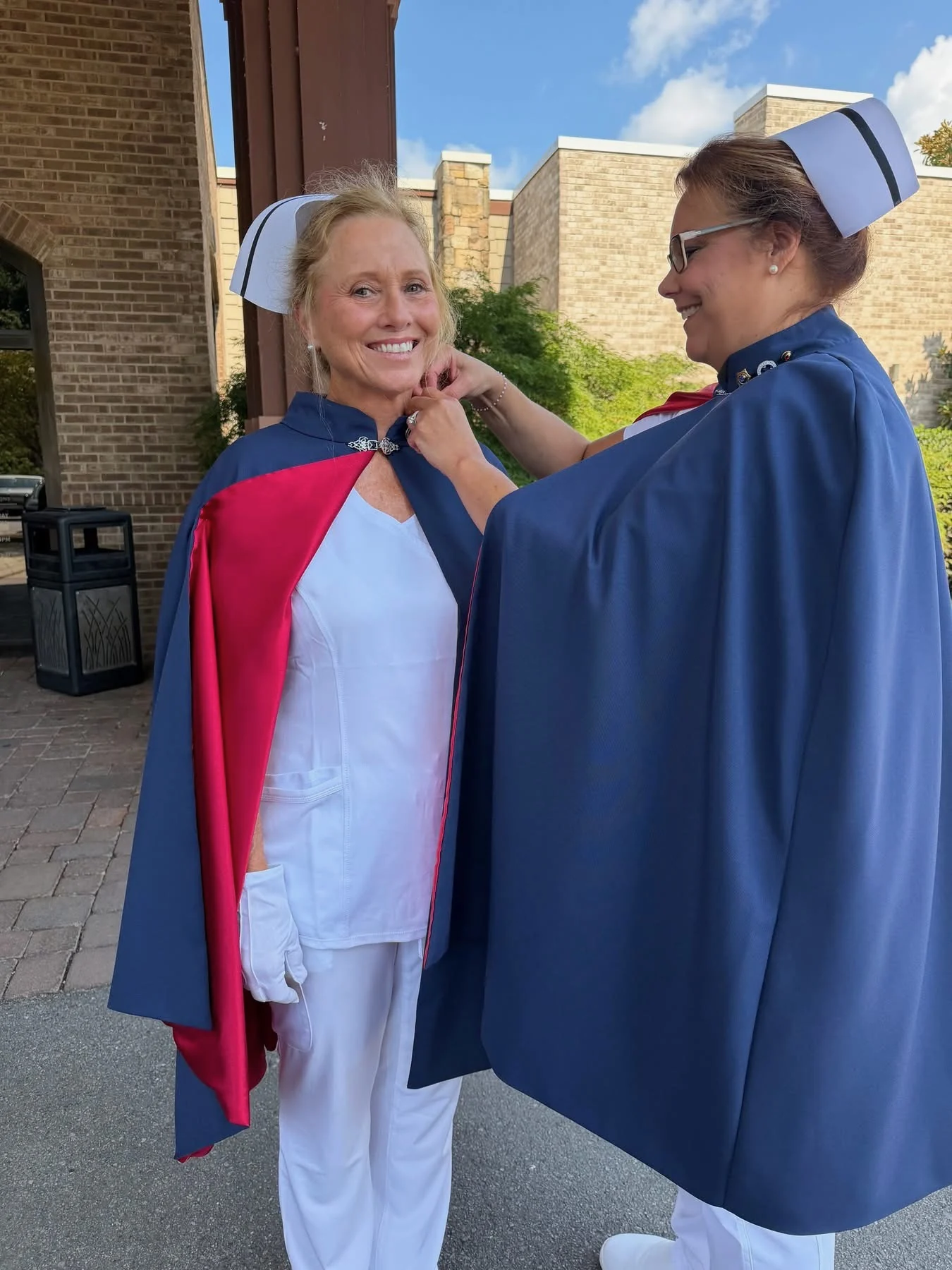 Kaye Ball in a white uniform, banded nurse’s cap and red-lined navy cape, smiling as Christina Trentham fastens her first-service pin at her collar outside a brick building.