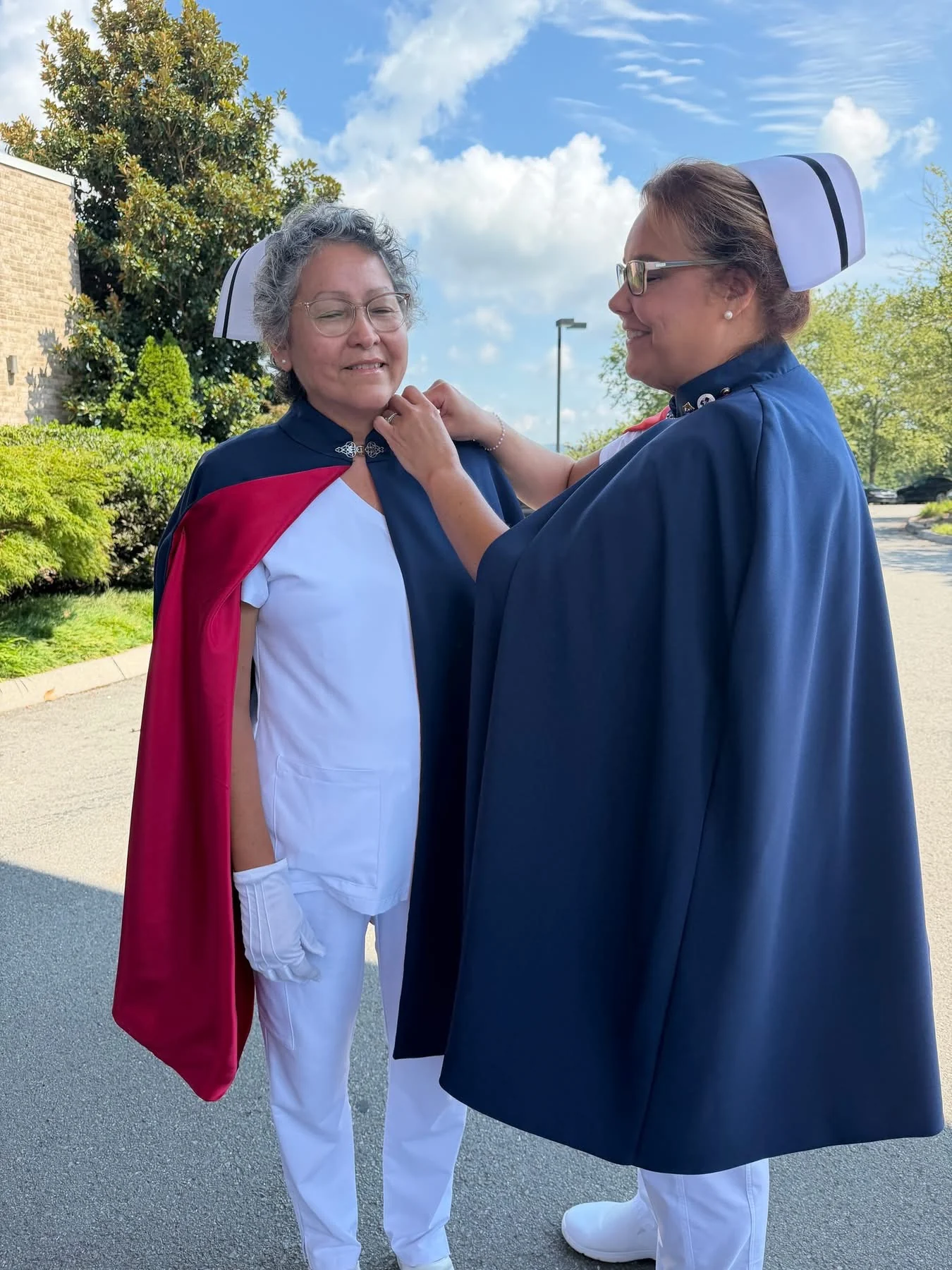 Patty Taylor in a white uniform, banded nurse’s cap and red-lined navy cape, smiling as Christina Trentham fastens her first-service pin at her collar under a blue sky.