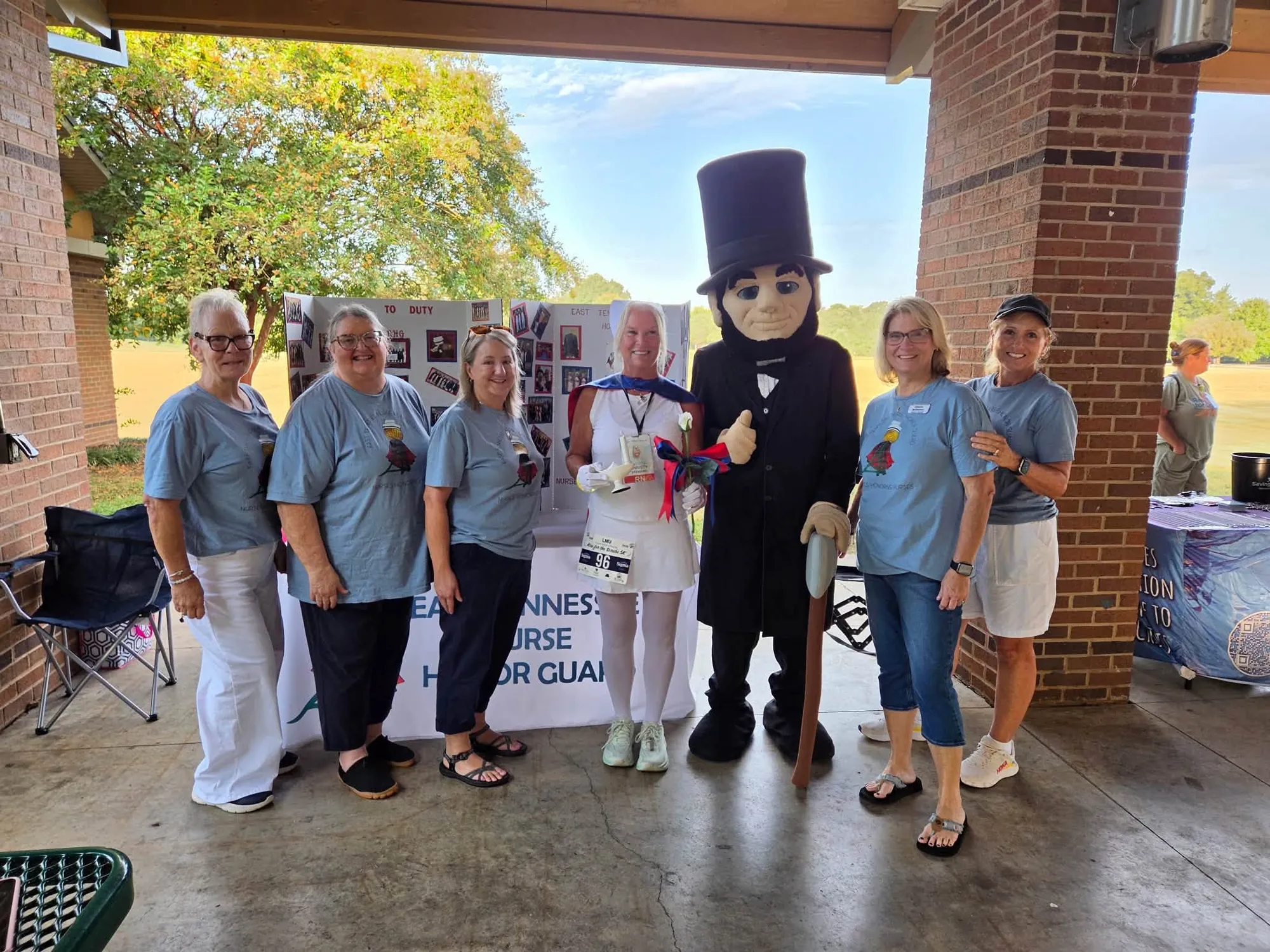 Five East Tennessee Nurse Honor Guard members in the chapter's light blue T-shirts under a park pavilion, with a runner dressed in white with a blue cape lined in red, holding a Nightingale lamp and a white rose, and a costumed Abraham Lincoln mascot giving a thumbs-up, in front of the chapter's photo display board and banner.
