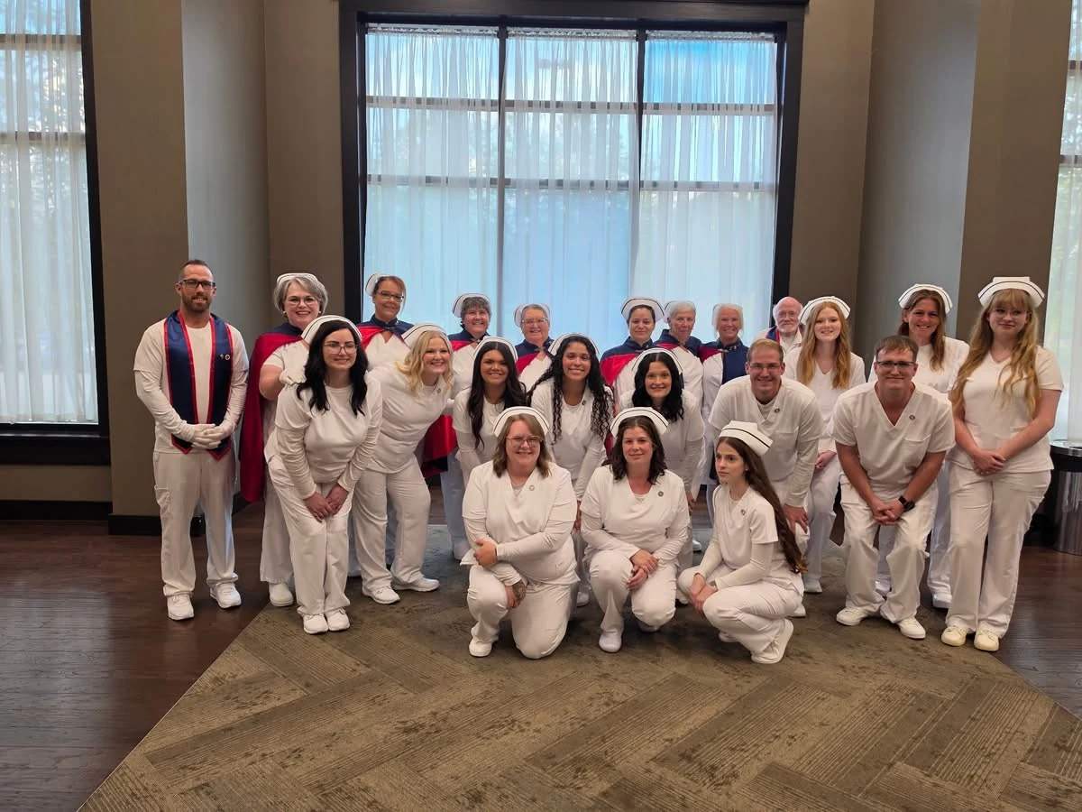The TCAT Harriman nursing class of 2026 in white uniforms and caps, three of them kneeling at the front, with East Tennessee Nurse Honor Guard members in navy and red capes standing behind them at their pinning ceremony on 27 August 2026.