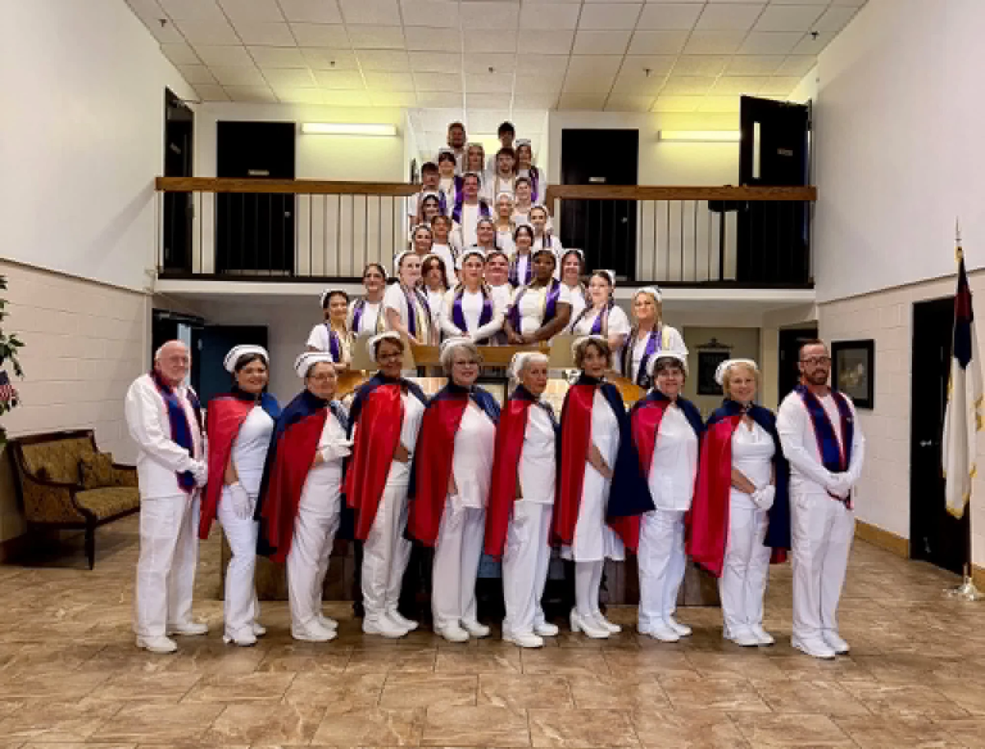 East Tennessee Nurse Honor Guard members in white uniforms with navy and red capes standing in a line across the front of a hall, with the TCAT Jacksboro nursing class of 2026 in white uniforms and purple-and-white stoles arranged on the staircase behind them on 28 August 2026.