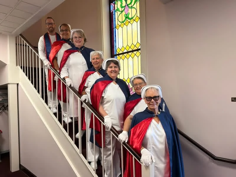Seven East Tennessee Nurse Honor Guard members in white uniforms, white caps and navy capes with red linings, standing in a line down a staircase beside a stained-glass window at the TCAT Oneida class of 2026 graduation.