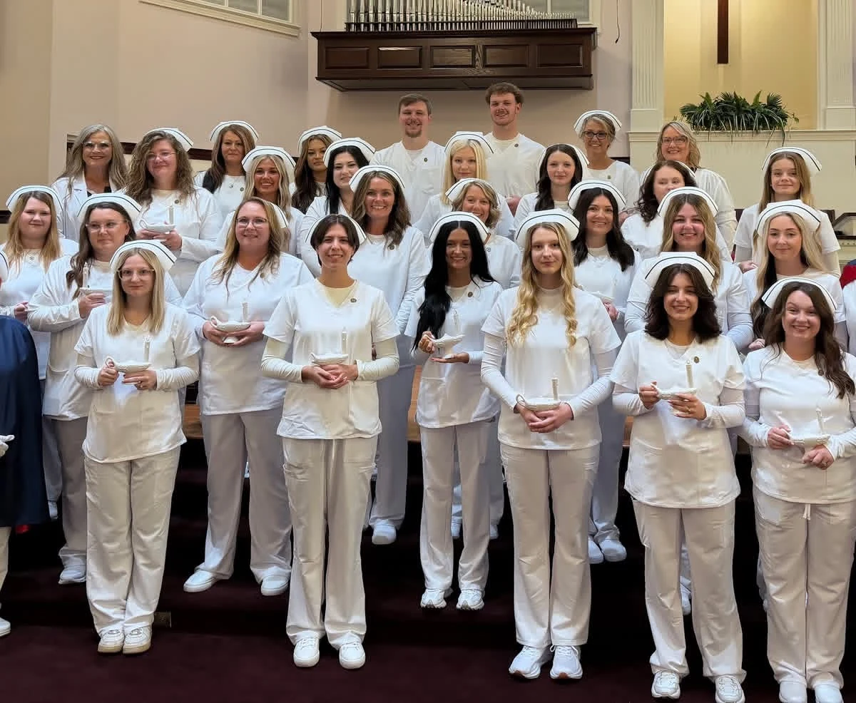 The TCAT Oneida nursing class of 2026 standing in rows in a chapel beneath an organ, all in white uniforms and nurses' caps, each holding a small white Nightingale lamp, at their graduation on 29 August 2026.