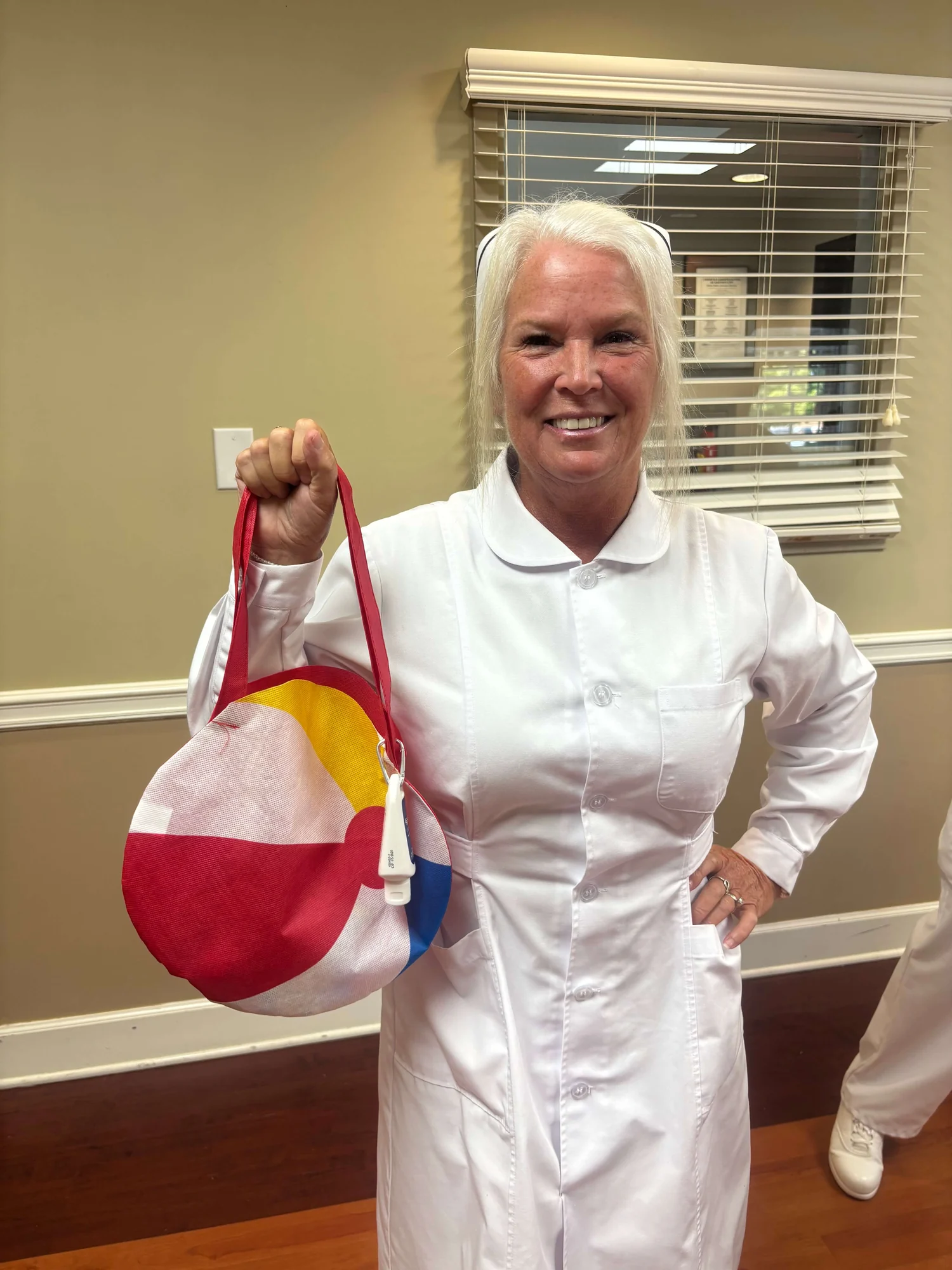 Cathy Felker in the chapter's white uniform, smiling and holding up one of the fabric treat bags she brings for members